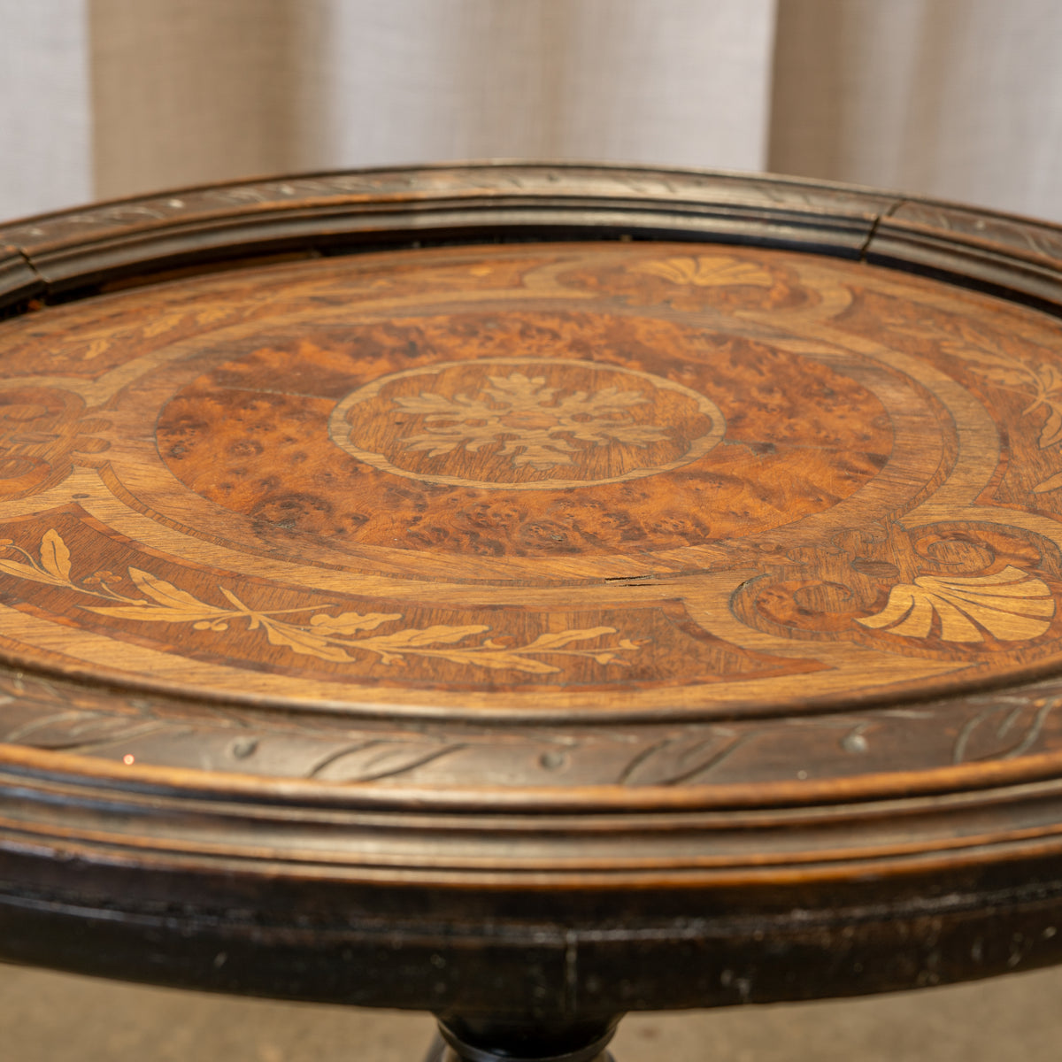 Close-up of an ornate round wooden table top with a detailed inlay pattern in warm brown and golden tones, surrounded by a dark carved wooden rim.