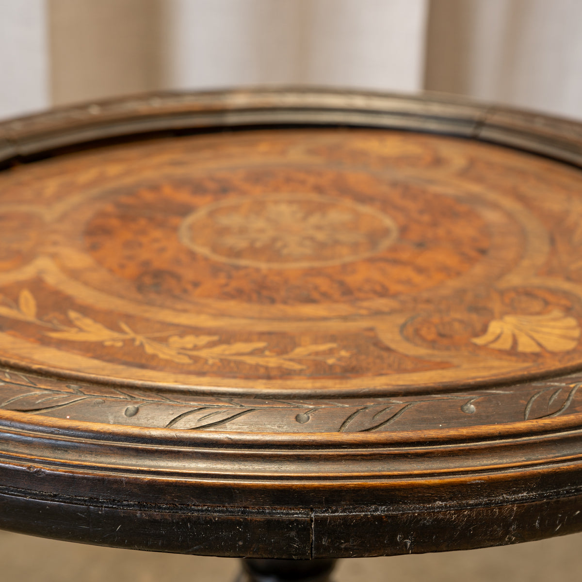 Close-up of an antique round wooden table top with an inlaid floral pattern and carved decorative border, showing warm brown wood tones and dark metal edging.