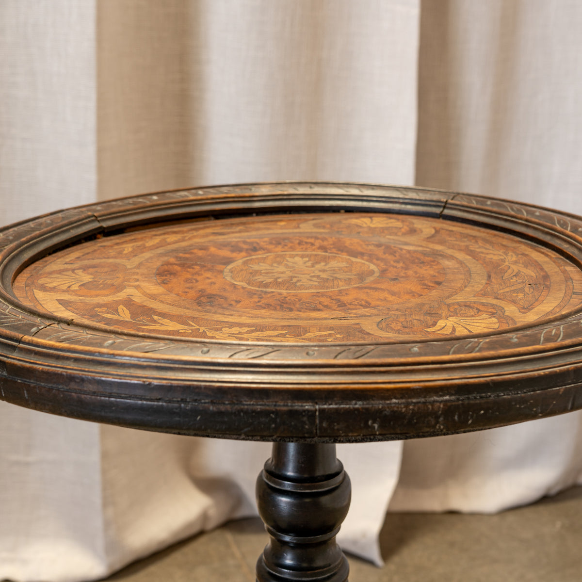 An antique round wooden table with a dark carved frame and a detailed inlay top featuring warm brown and golden tones, set against a neutral draped fabric backdrop.