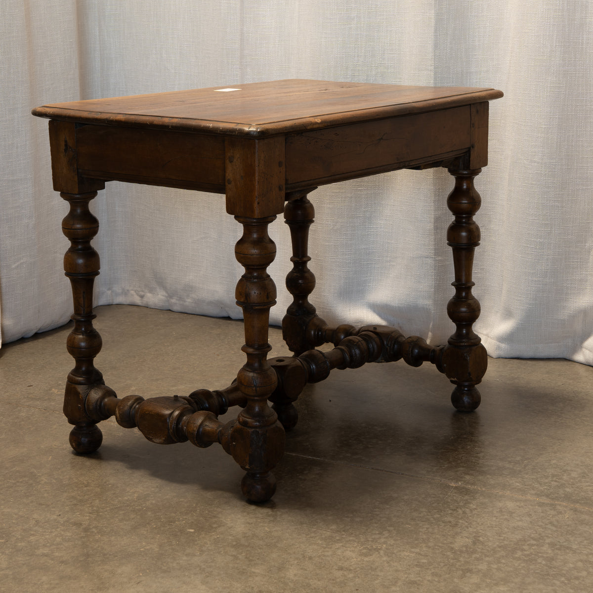 An antique dark brown wooden table with turned spindle legs and a decorative stretcher bar, set against a draped white fabric backdrop.