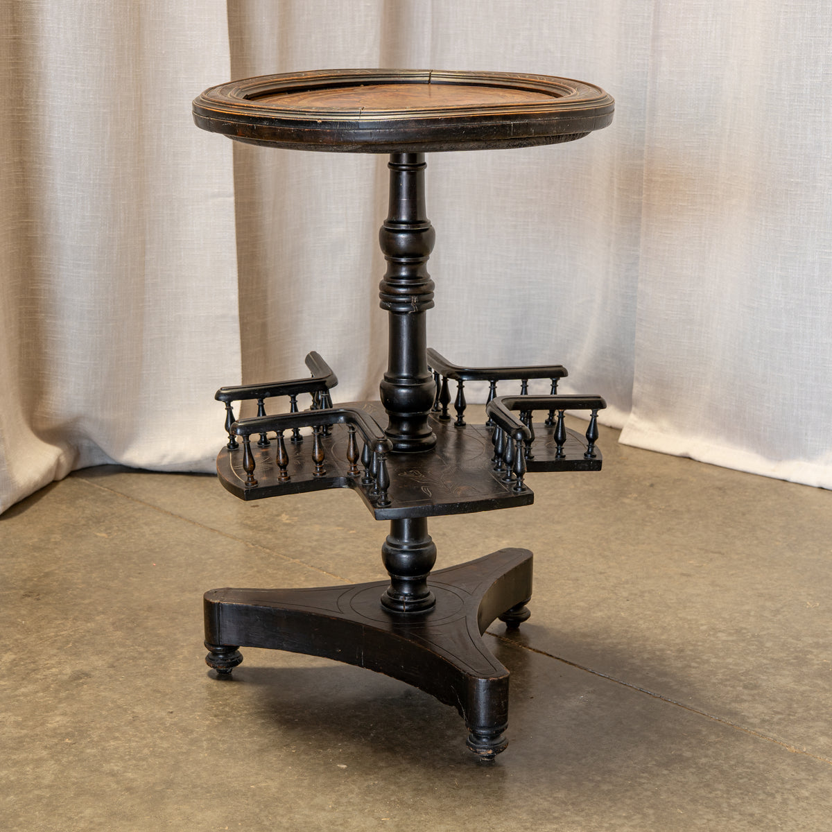 A vintage black wooden bar cart with a round top, turned central pedestal, and three tiered side shelves, photographed against a neutral draped fabric backdrop.
