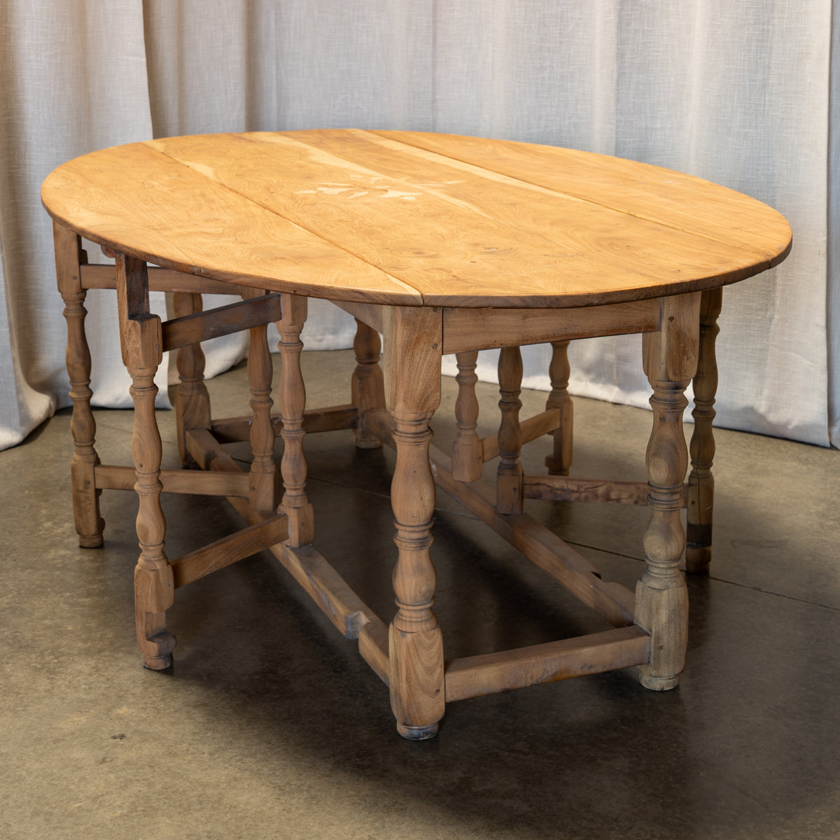 An oval wooden dining table with turned spindle legs and stretchers, shown in a rustic setting with draped fabric curtains and a concrete floor.