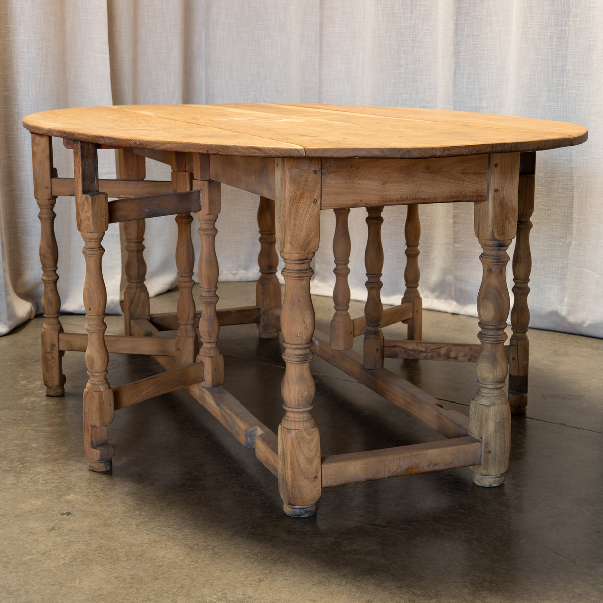 An antique-style wooden dining table with a round natural wood top and turned spindle legs, set against a neutral fabric backdrop.