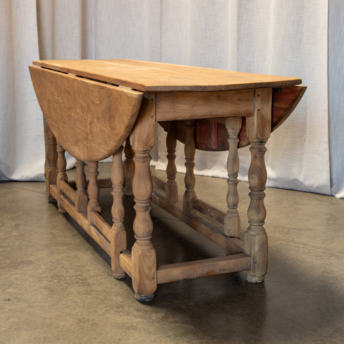 A rustic wooden drop-leaf table with turned spindle legs and a light natural wood finish, photographed indoors against a draped fabric backdrop.