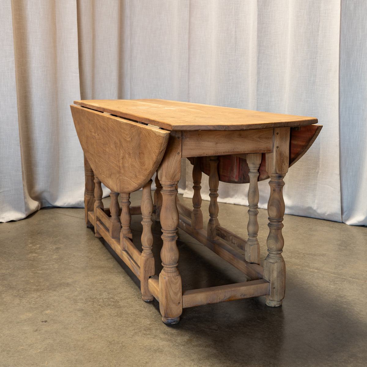 A rustic wooden drop-leaf table with turned spindle legs and a natural oak finish, shown with one leaf folded down, set against a neutral draped fabric backdrop.