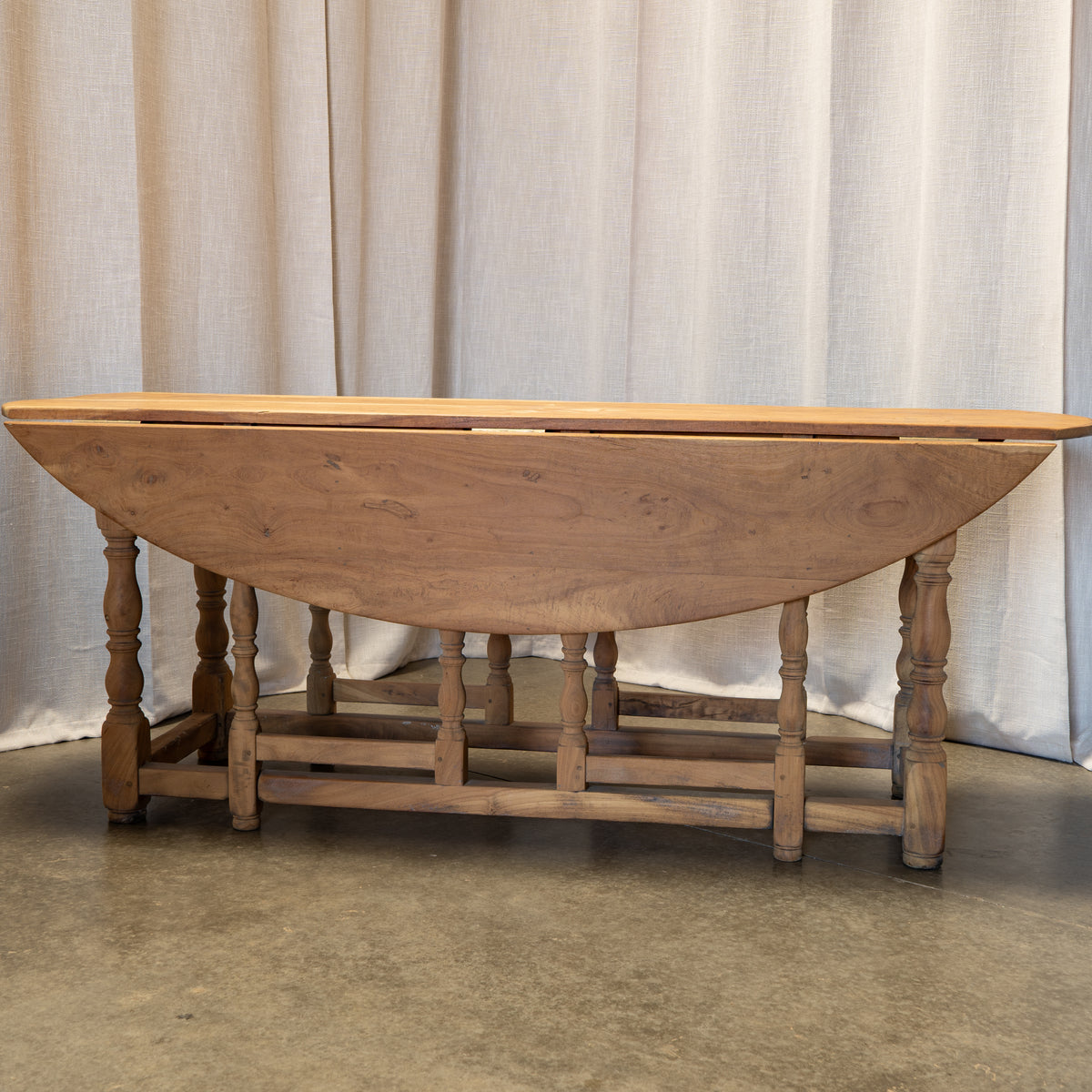 A rustic wooden drop-leaf table with turned spindle legs and a lower stretcher bar, shown against a neutral beige curtain backdrop.