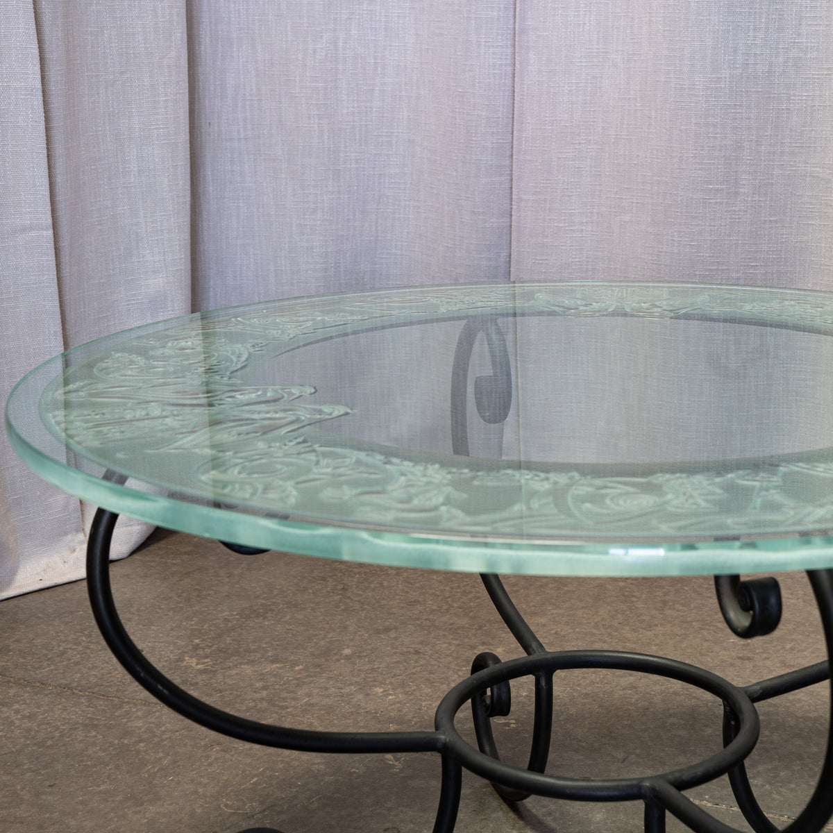 Round glass-top table with a black metal scrollwork base, photographed indoors against a light gray curtain backdrop.