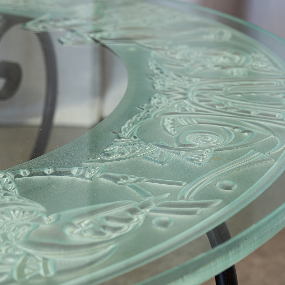 Close-up of a round glass tabletop with a frosted, etched decorative pattern featuring stylized figures and scrollwork in a pale mint-green tint.