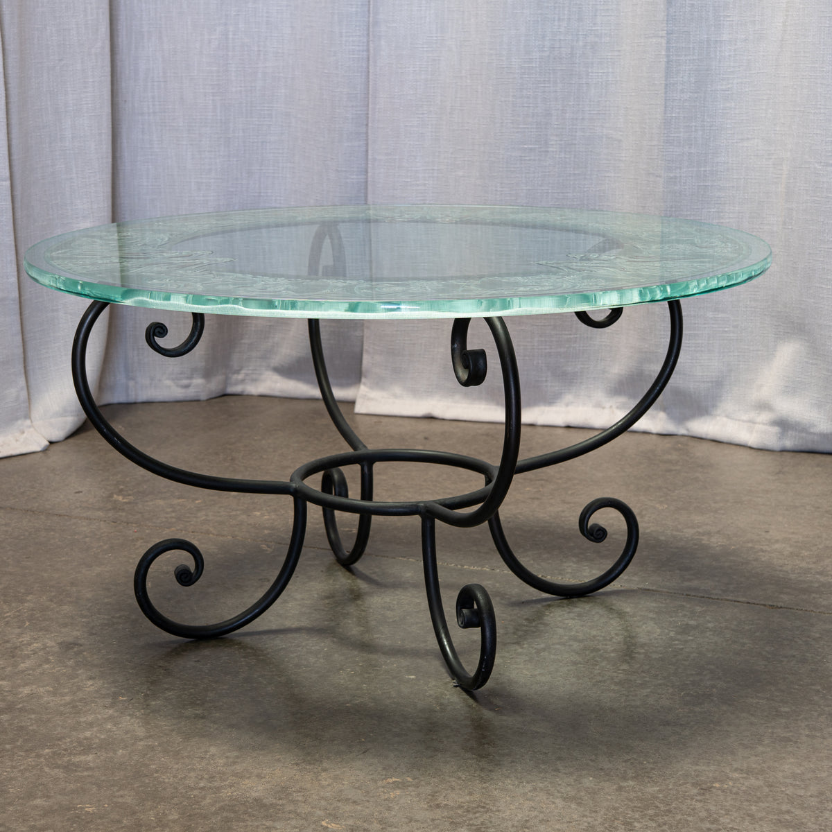 A round glass-top coffee table with a black wrought iron scrollwork base, set against a backdrop of light gray fabric drape and concrete floor.