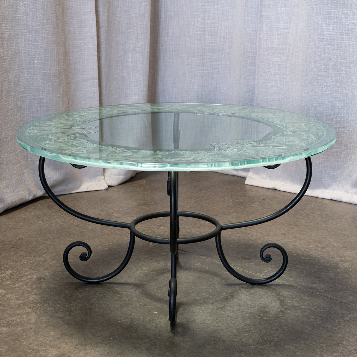 Round outdoor coffee table with a clear glass top featuring a light green etched floral border and a black wrought iron scroll base, set against a neutral draped fabric backdrop.