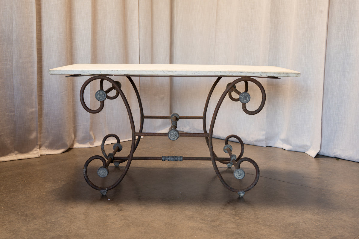 A rustic console table featuring a distressed white-painted stone top and an ornate dark brown wrought iron base with scrollwork and cast metal accents, set against a neutral draped fabric backdrop.