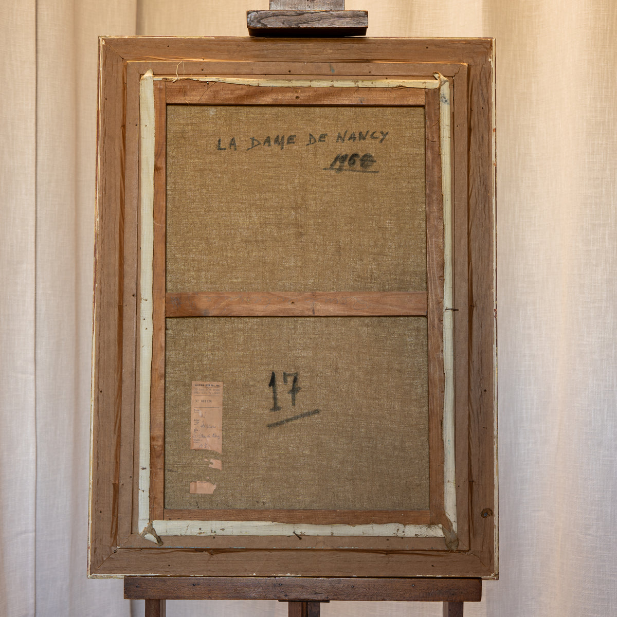 The back of a framed canvas print on an easel, showing the linen canvas, wooden frame, and handwritten text 'LA DAME DE NANCY' and 'Mme' on a beige curtain background.