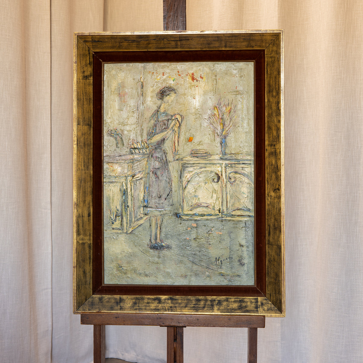 A framed vintage painting depicting a woman in a domestic interior, mounted on a wooden easel against a draped beige fabric backdrop.