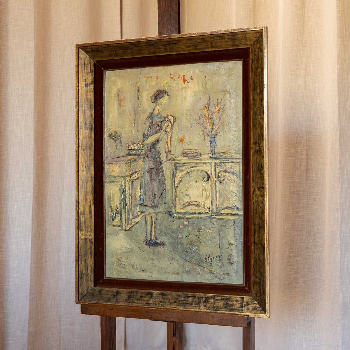 A framed painting on an easel, depicting a woman standing in a room with furniture and a vase. The artwork is housed in a distressed, ornate gold and brown frame against a beige curtain background.