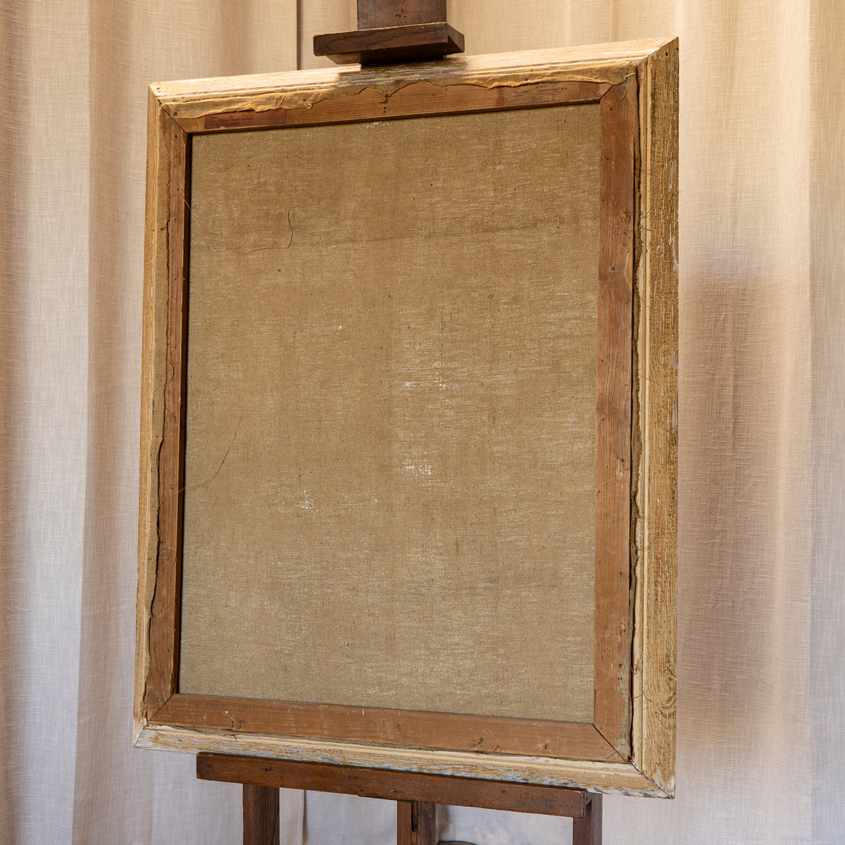 An antique wooden picture frame with a textured beige linen backing, displayed on a wooden easel against a neutral beige curtain background.