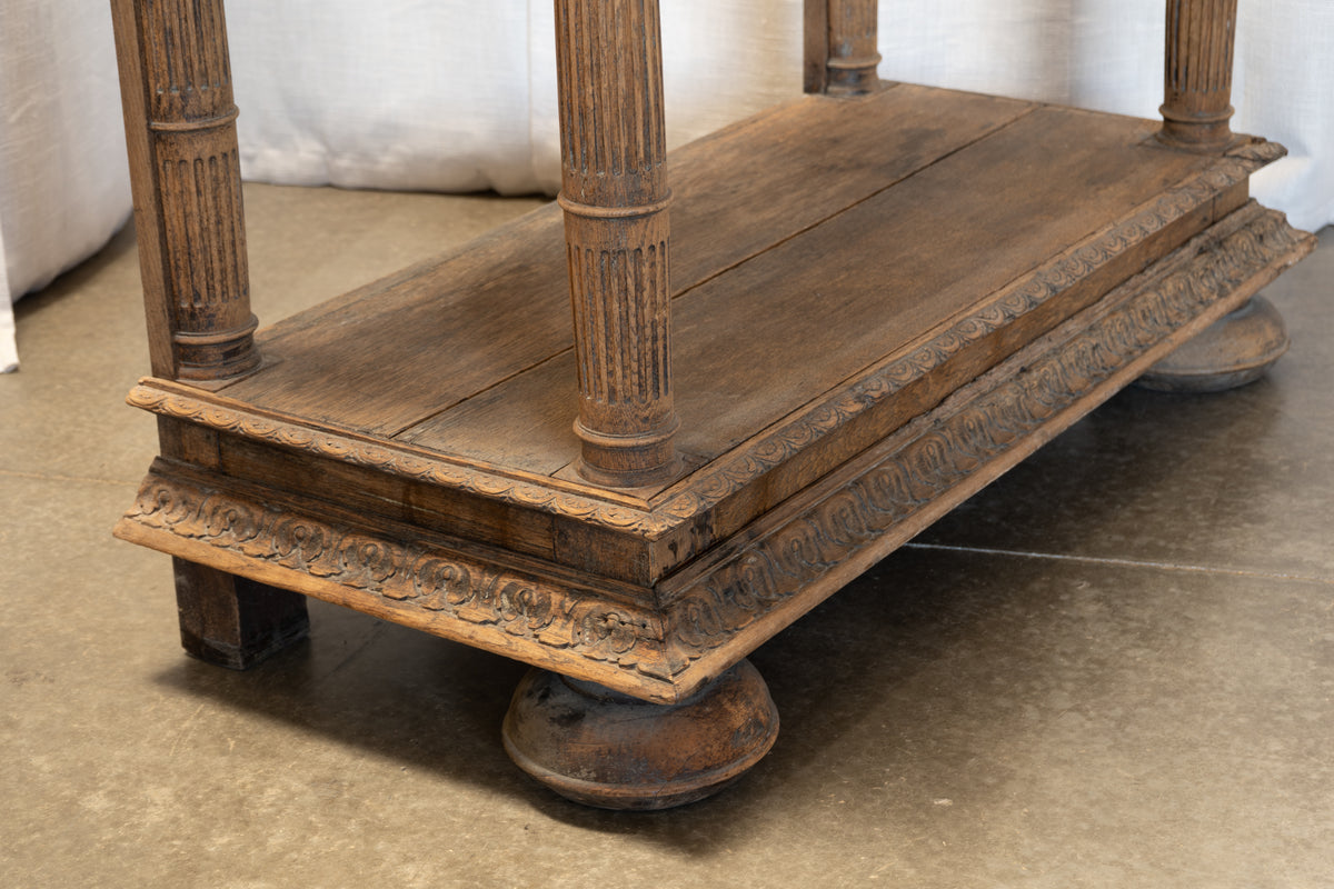 A close-up of a dark brown carved wooden stand or low shelf with fluted legs, photographed indoors on a concrete floor.