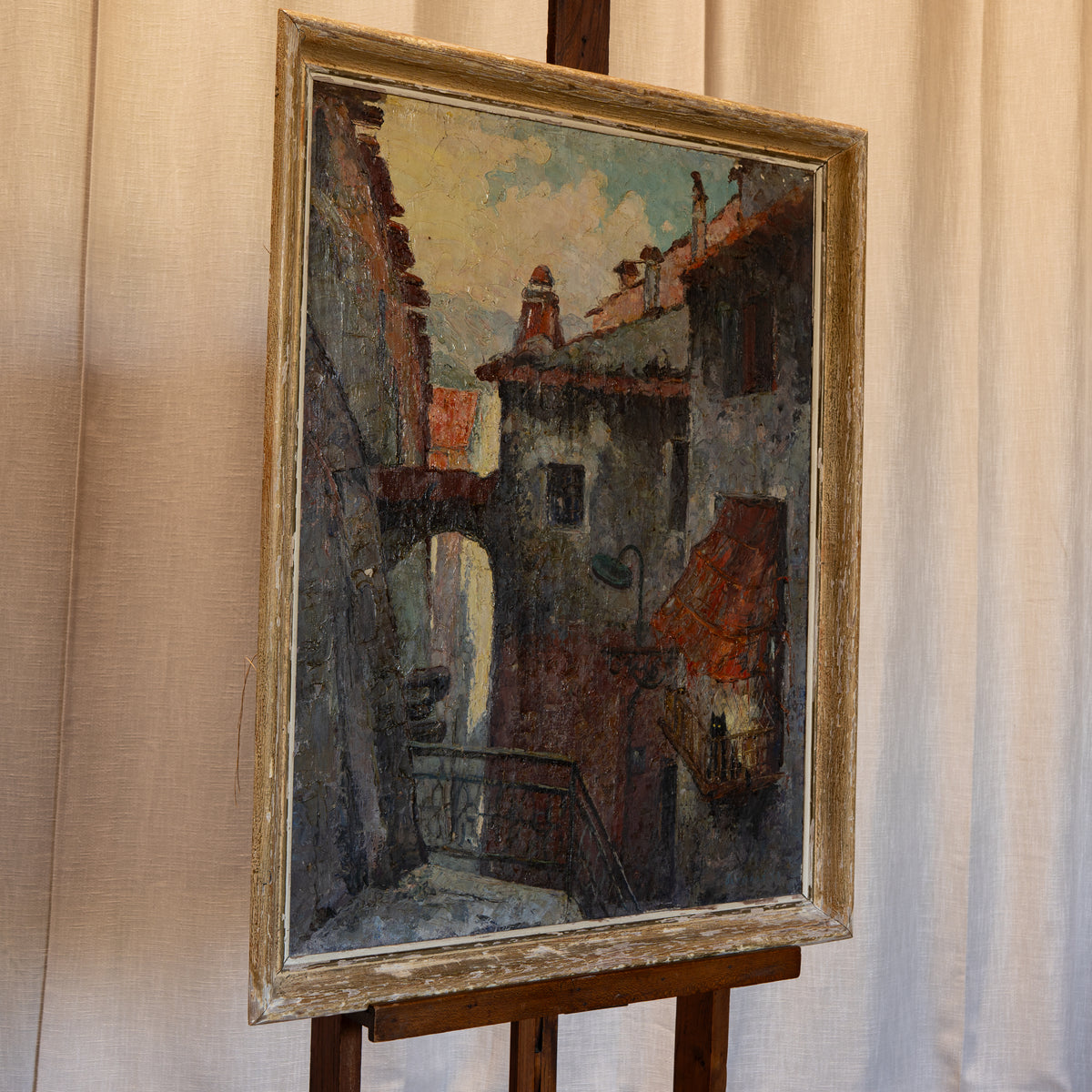 A framed oil painting depicting a narrow European alleyway with rustic buildings, rendered in muted earth tones of gray, brown, and terracotta, mounted on a wooden easel against a draped beige fabric backdrop.
