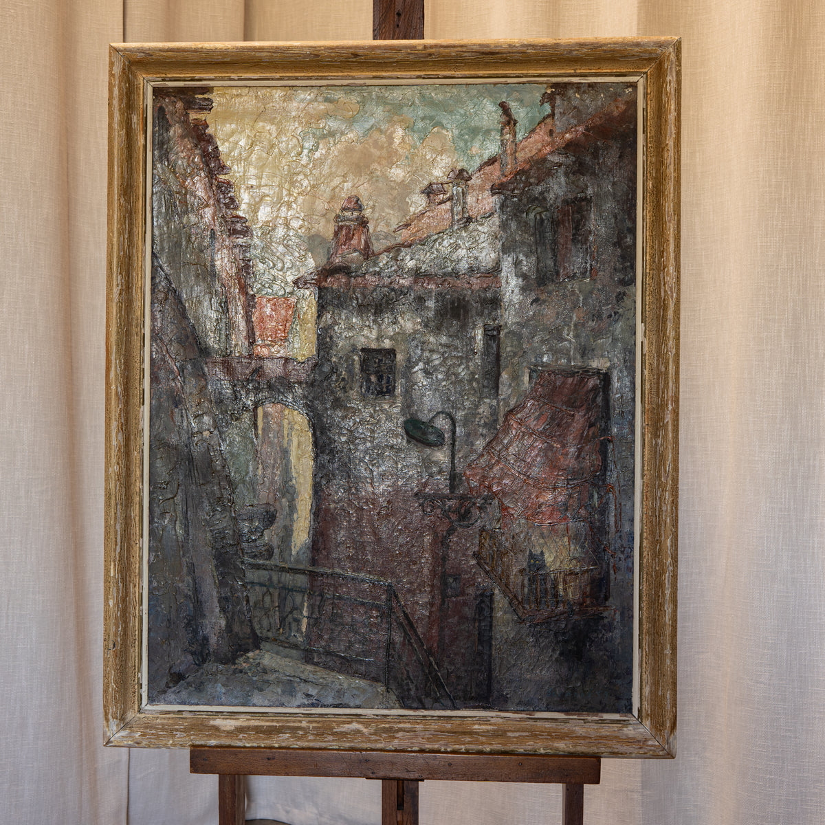 An oil painting of a narrow European alleyway with textured brushstrokes in gray, brown, red, and beige tones, housed in a distressed gold frame and displayed on a wooden easel against a draped beige fabric backdrop.