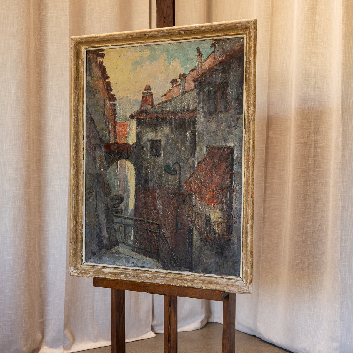 A framed oil painting of a European alleyway with rustic buildings, displayed on a wooden easel in front of beige curtains.