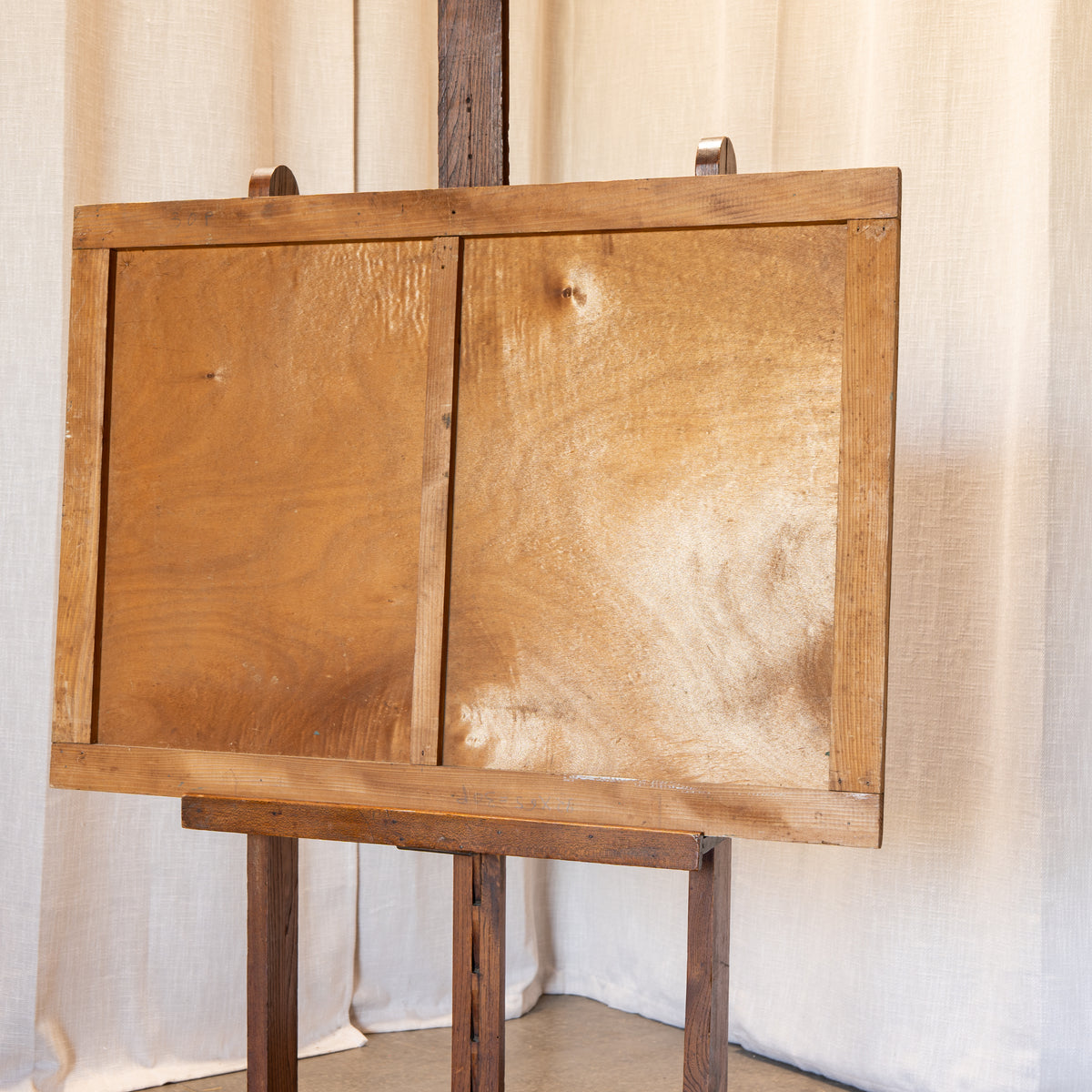 A rustic wooden cabinet or panel with two doors, displayed on a dark wooden easel against a draped cream fabric backdrop.