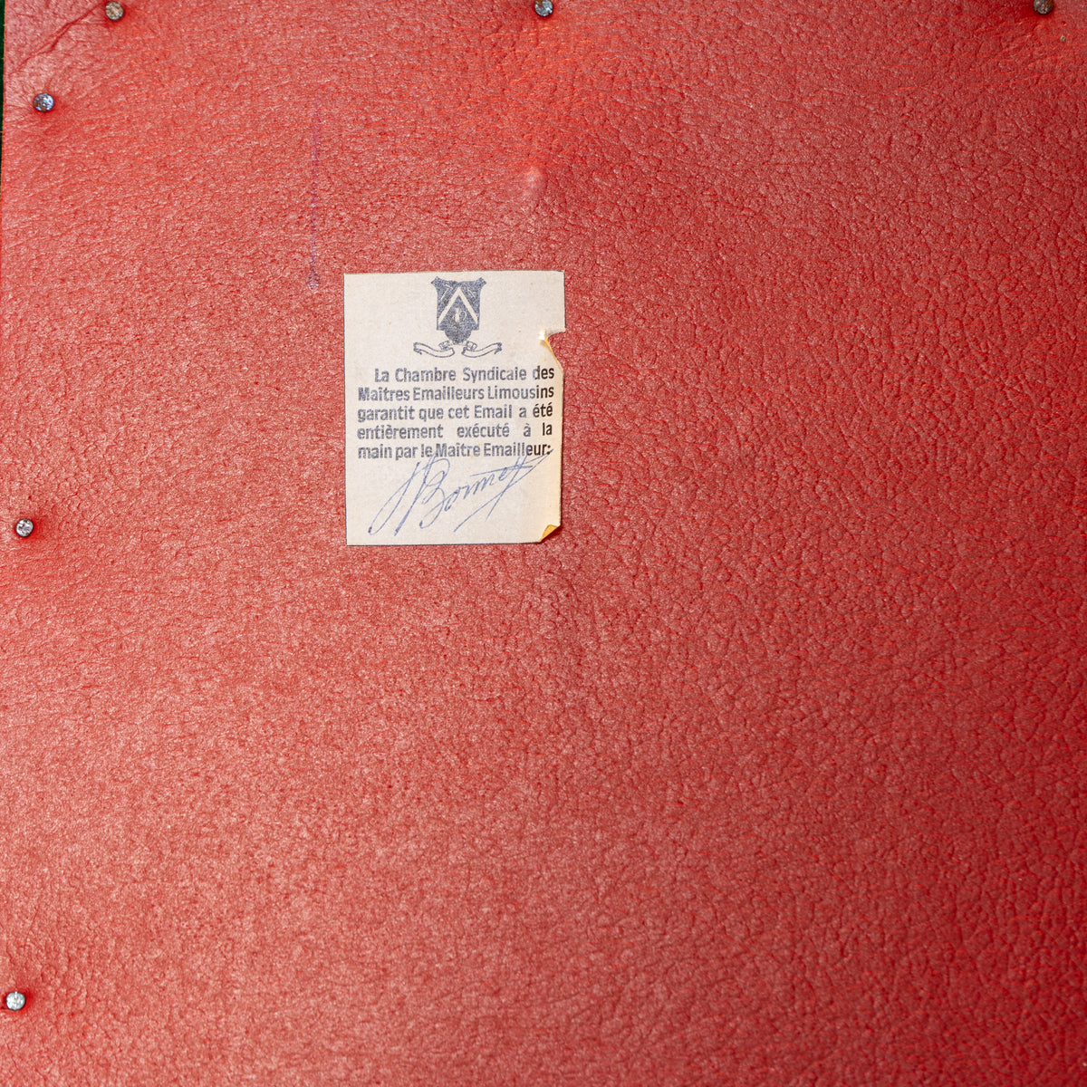 A close-up of a textured red surface with a small beige paper tag attached near the center, featuring a crest and handwritten text in blue ink.