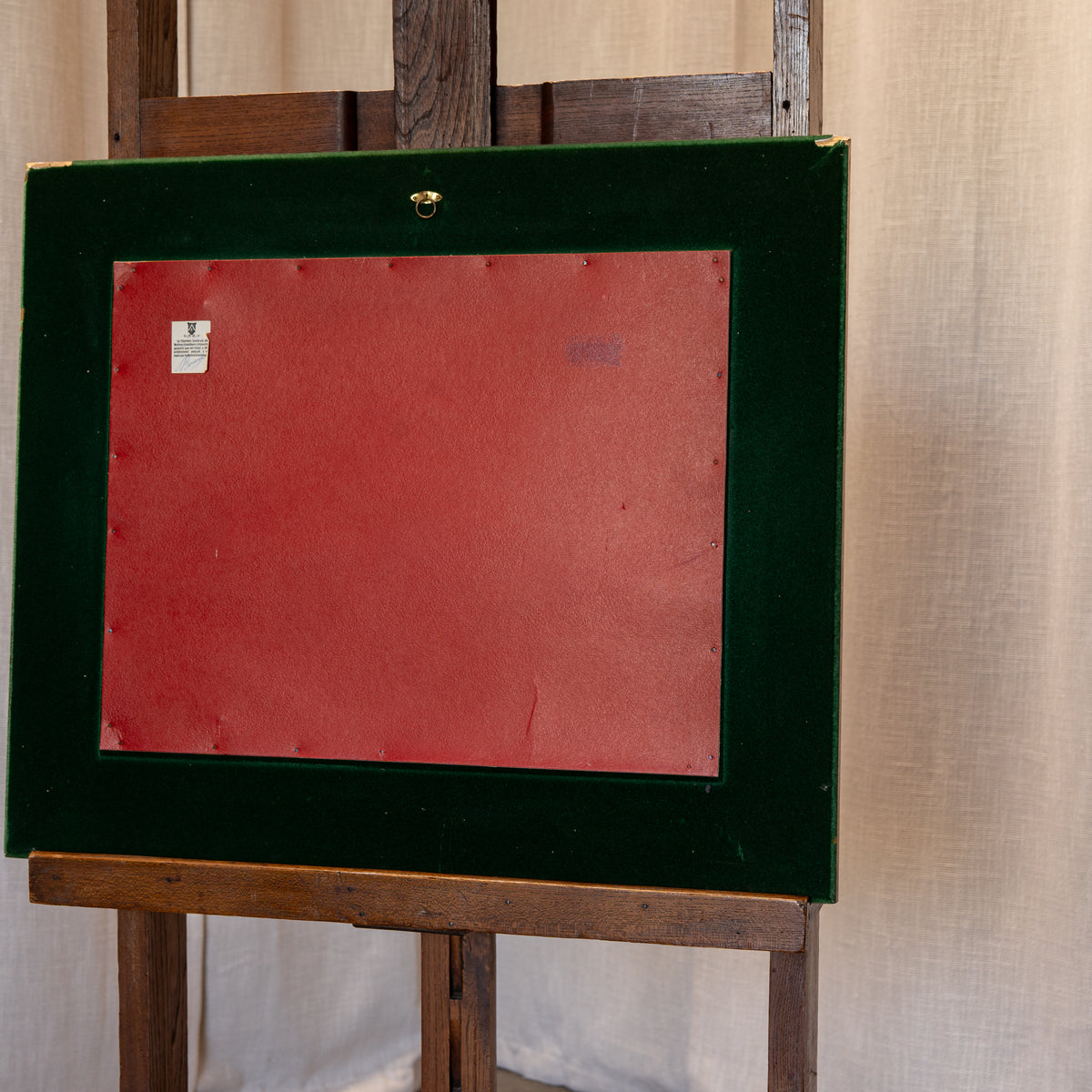 The back of a vintage picture frame with a dark green velvet border and a red backing board, displayed on a wooden easel against a beige curtain backdrop.