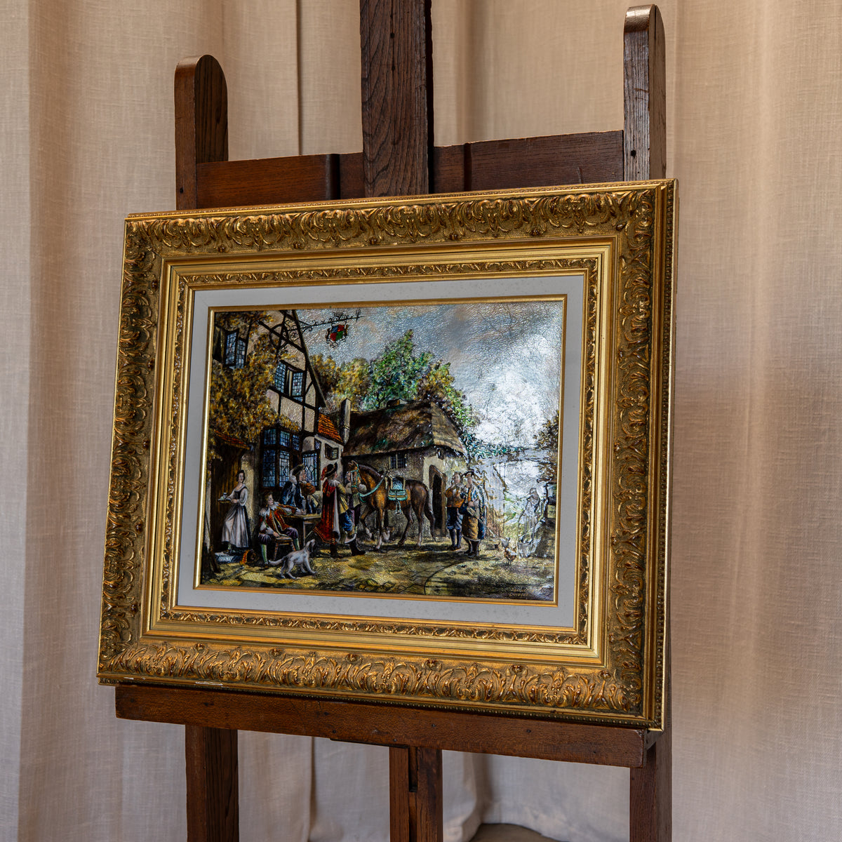 An ornate gold-framed painting depicting a rustic village street scene with figures and horses, displayed on a wooden easel against a beige curtain backdrop.