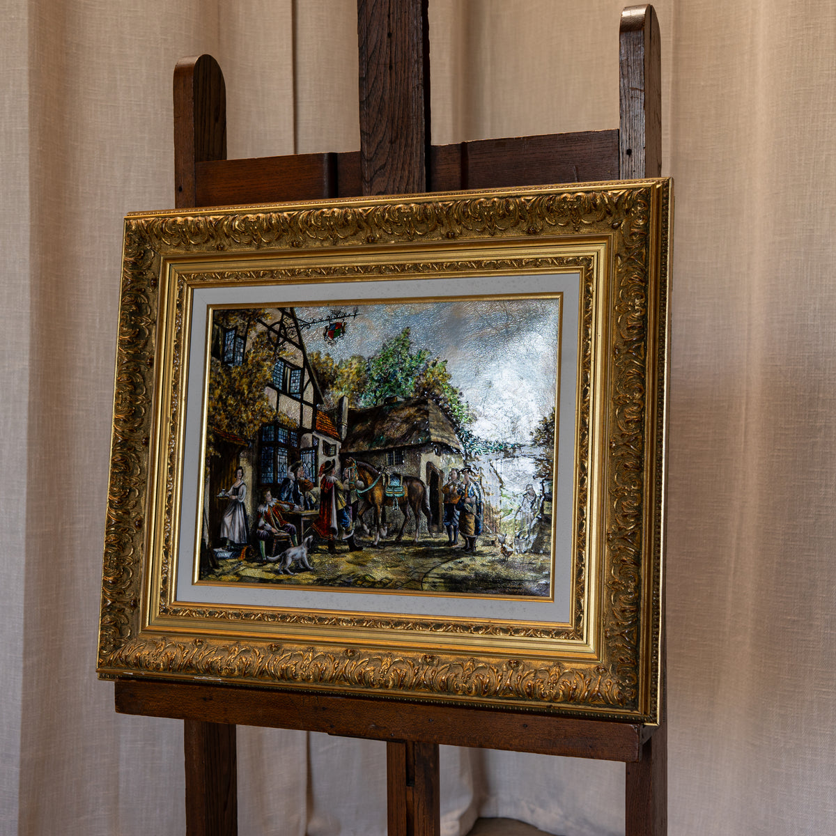 A framed painting depicting a rustic village street scene with figures and horses, set in an ornate gold frame and displayed on a wooden easel against a beige curtain backdrop.