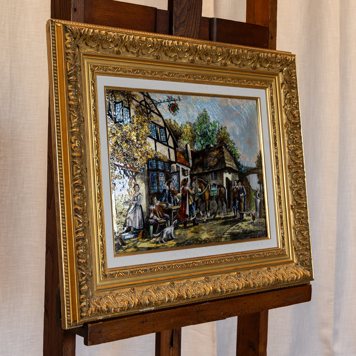 An ornate gold-framed painting depicting a historical village street scene with figures, trees, and buildings, displayed on a wooden easel against a light fabric backdrop.