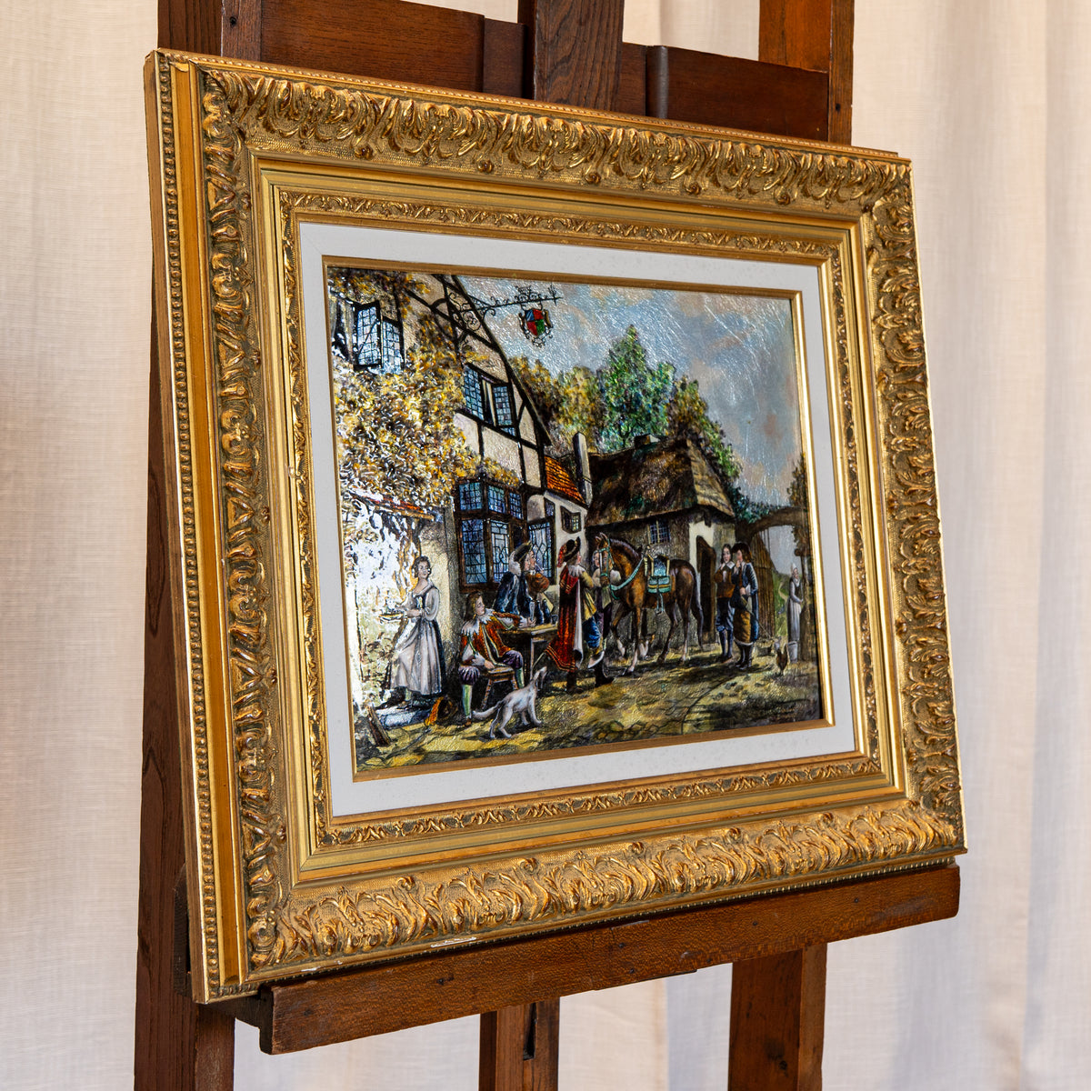 A framed painting depicting a rustic outdoor village scene with people gathered around tables by a tree, mounted in an ornate gold frame and displayed on a wooden easel against a light curtain background.