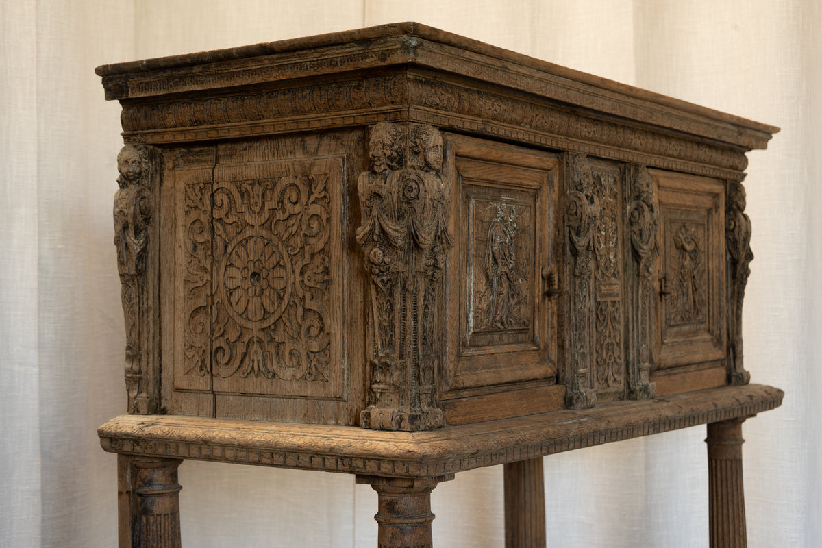 An antique carved wooden chest or cabinet on a matching pedestal stand, shown against a light fabric backdrop. The chest features intricate relief carvings, decorative figures, and a weathered dark brown wood finish.