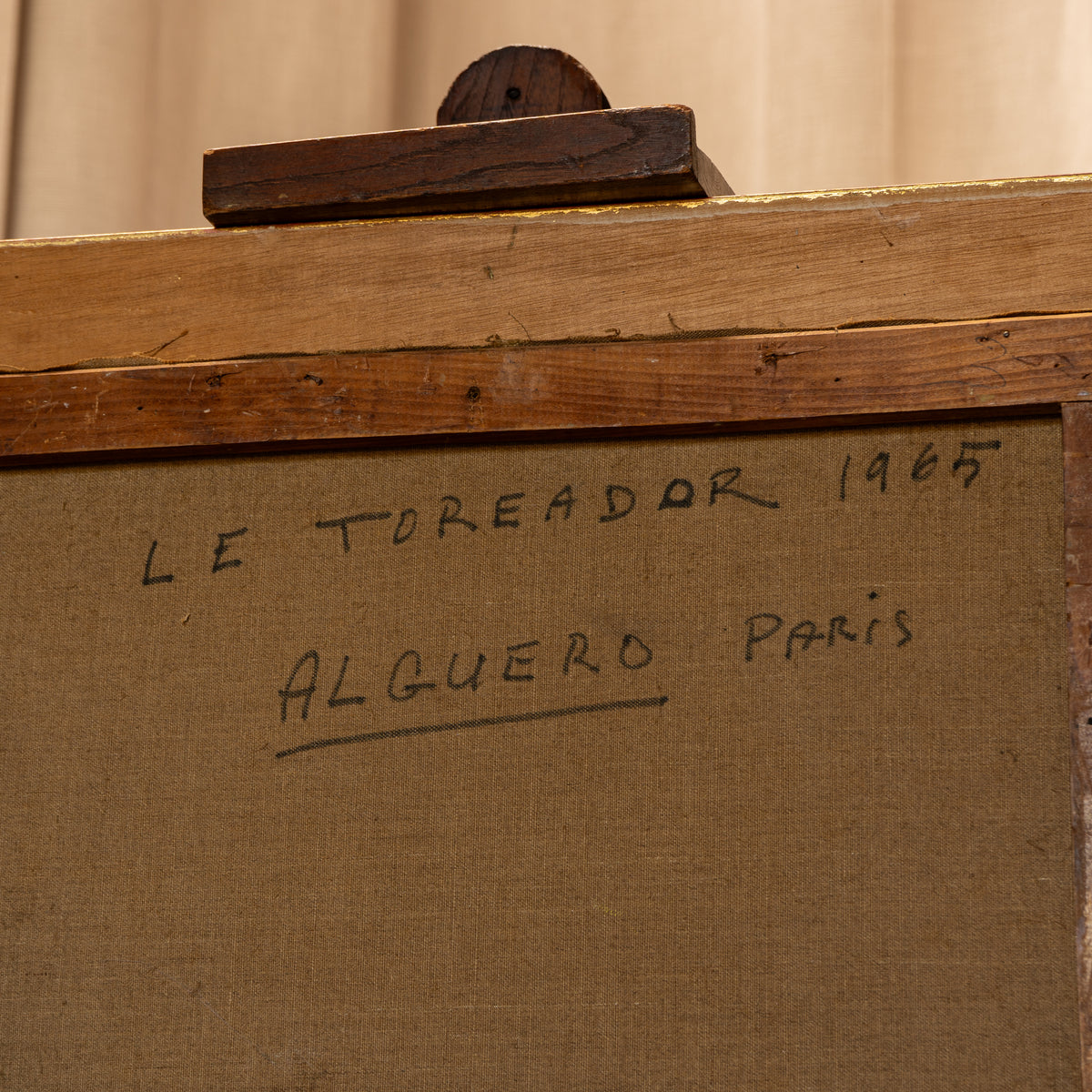 A close-up of a wooden framed canvas or print with handwritten text on the front reading 'LE TOREADOR 1965 ALGUERO Paris' on a tan fabric surface.
