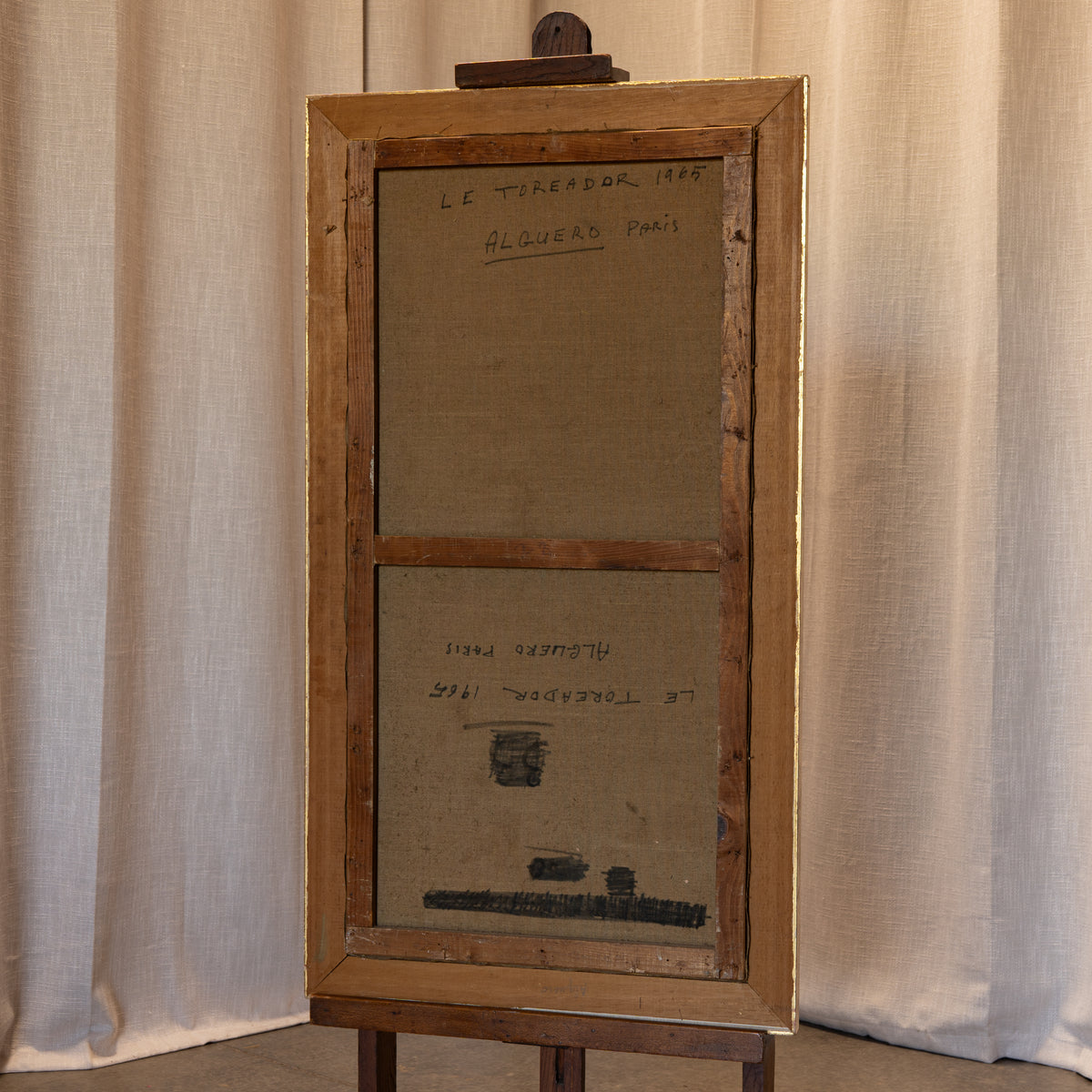 A framed textile artwork on an easel, featuring a rustic wooden frame with two panels of linen fabric in warm beige and brown tones, with handwritten text and small dark painted details. The background consists of draped beige fabric.