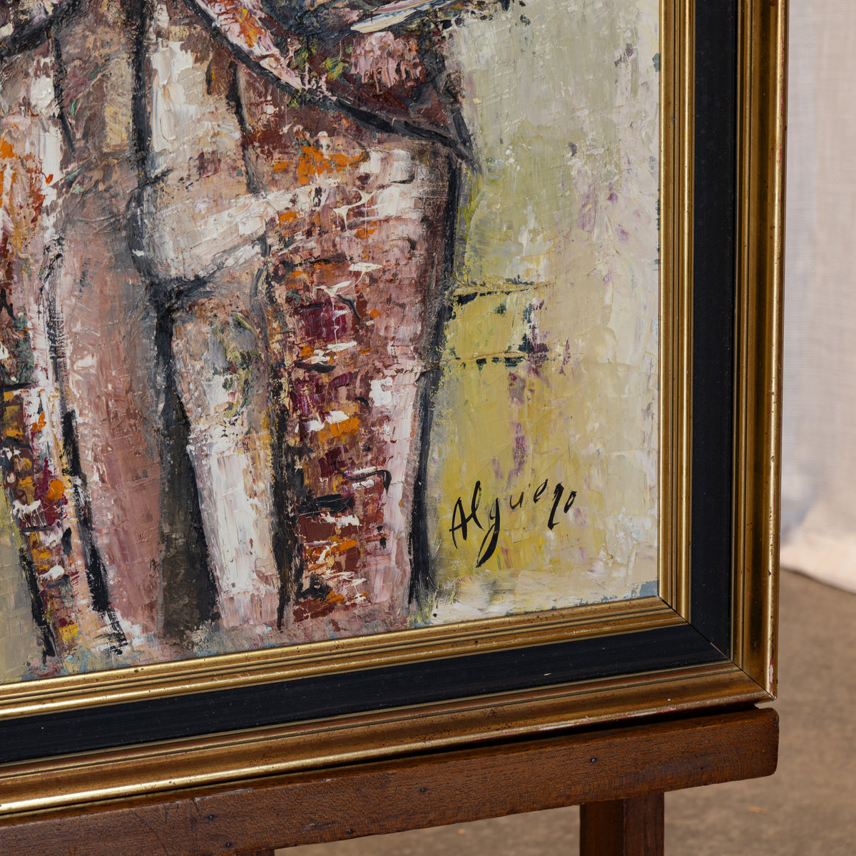 Close-up of a framed painting on an easel, showing textured brushstrokes in beige, brown, white, and orange tones with a gold frame and black mat.
