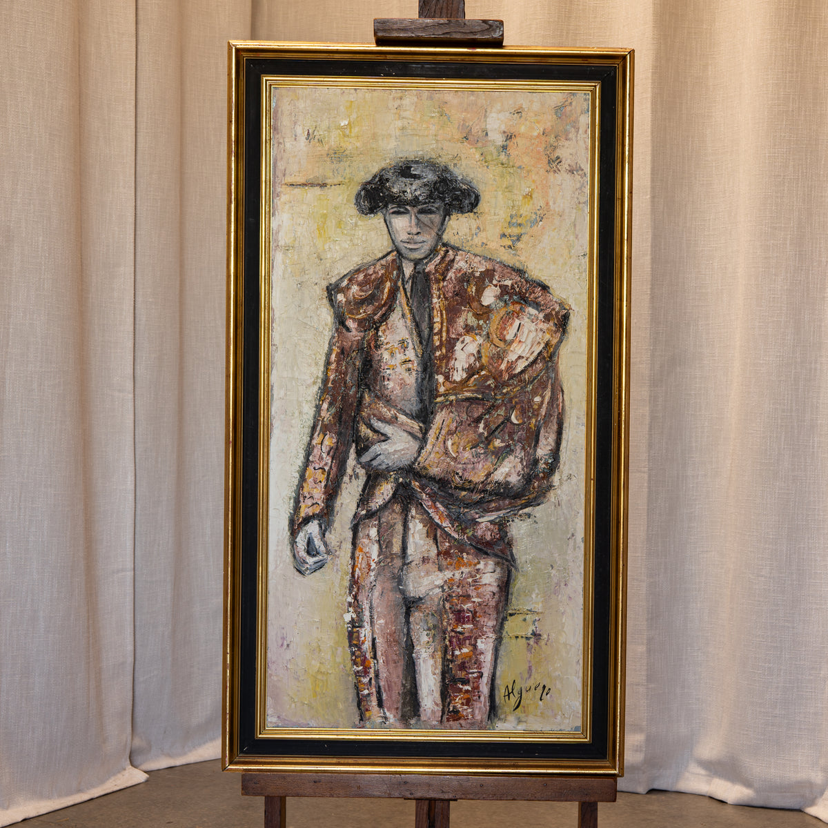 A framed painting of a bullfighter in a textured brown and beige suit, displayed on a wooden easel against a neutral curtain backdrop.