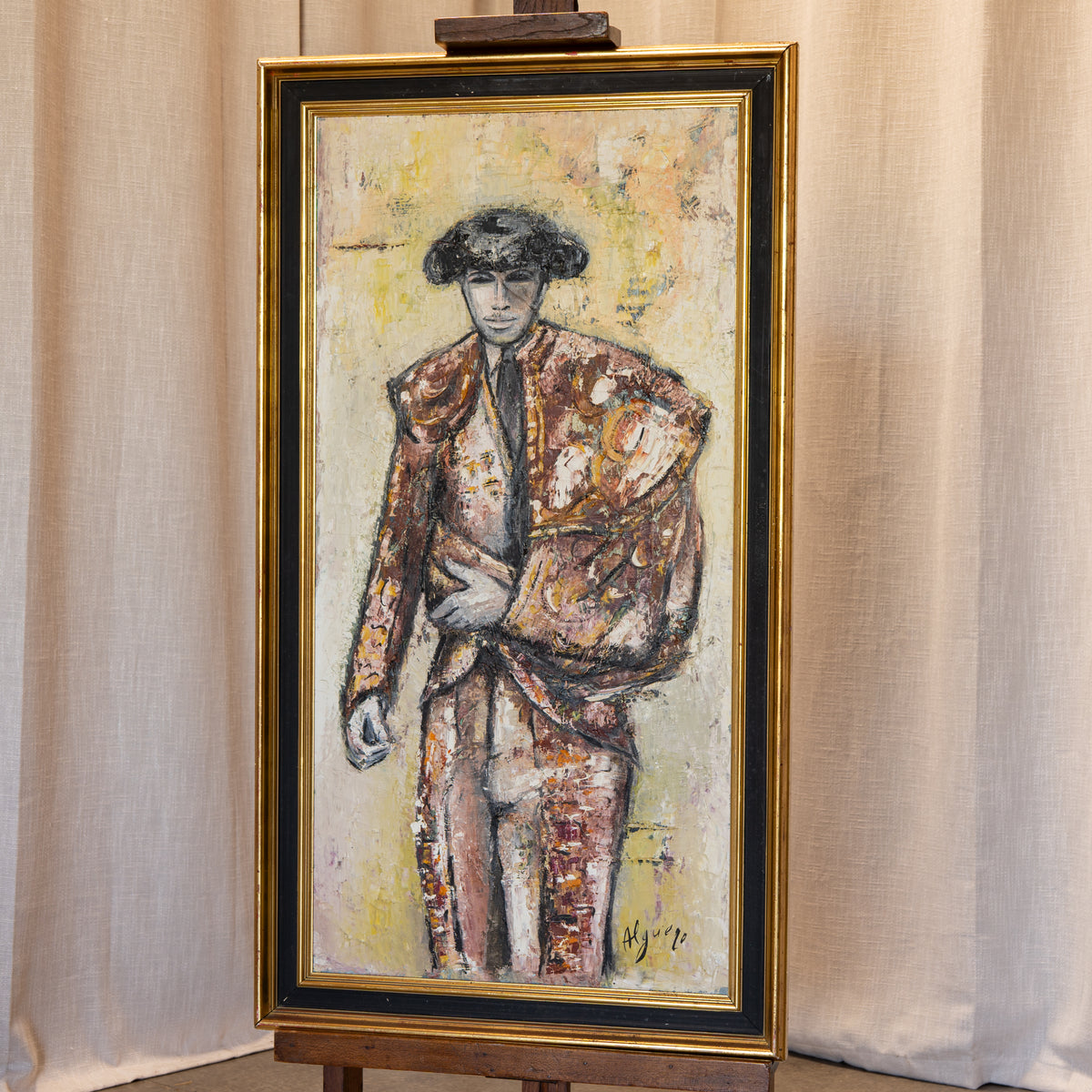 A framed vertical painting on an easel, depicting a stylized figure in a textured, painterly suit against a warm cream and yellow background. The painting is housed in a gold frame with a black inner border, set against a draped beige fabric backdrop.