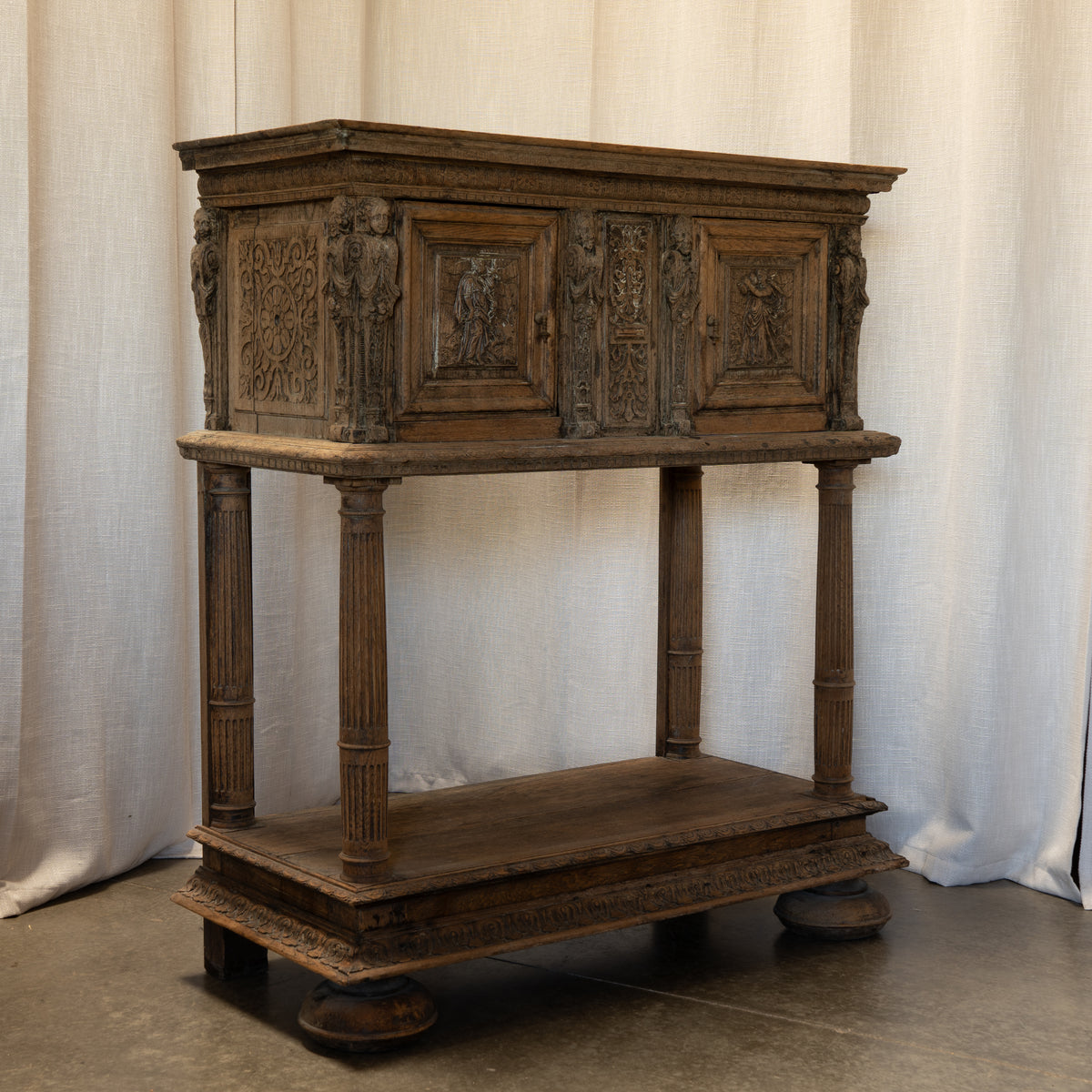 An ornate antique wooden cabinet or console table with carved doors, decorative side figures, four fluted legs, and a lower shelf, photographed against a draped white fabric backdrop.