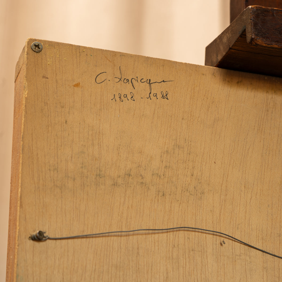 Close-up of the back of a light wood panel with handwritten text reading 'C. Dapicque 1928-1988' and a visible hanging wire, photographed indoors against a beige wall.