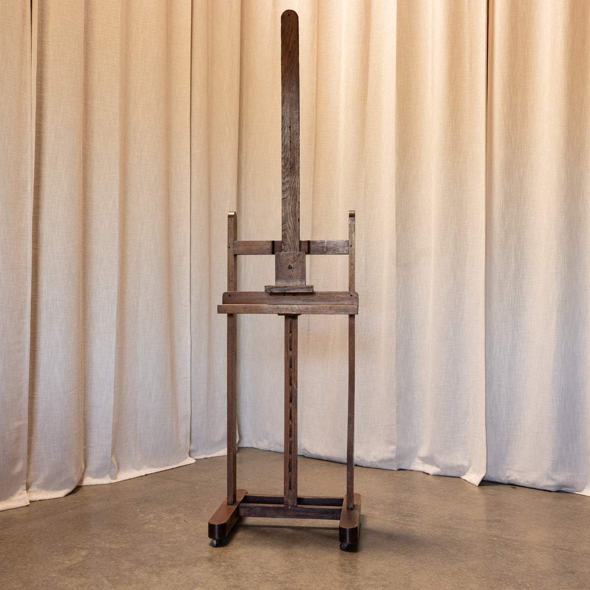 A dark brown wooden easel with a tall vertical arm and adjustable crossbar system, standing on a concrete floor in front of draped beige fabric curtains.