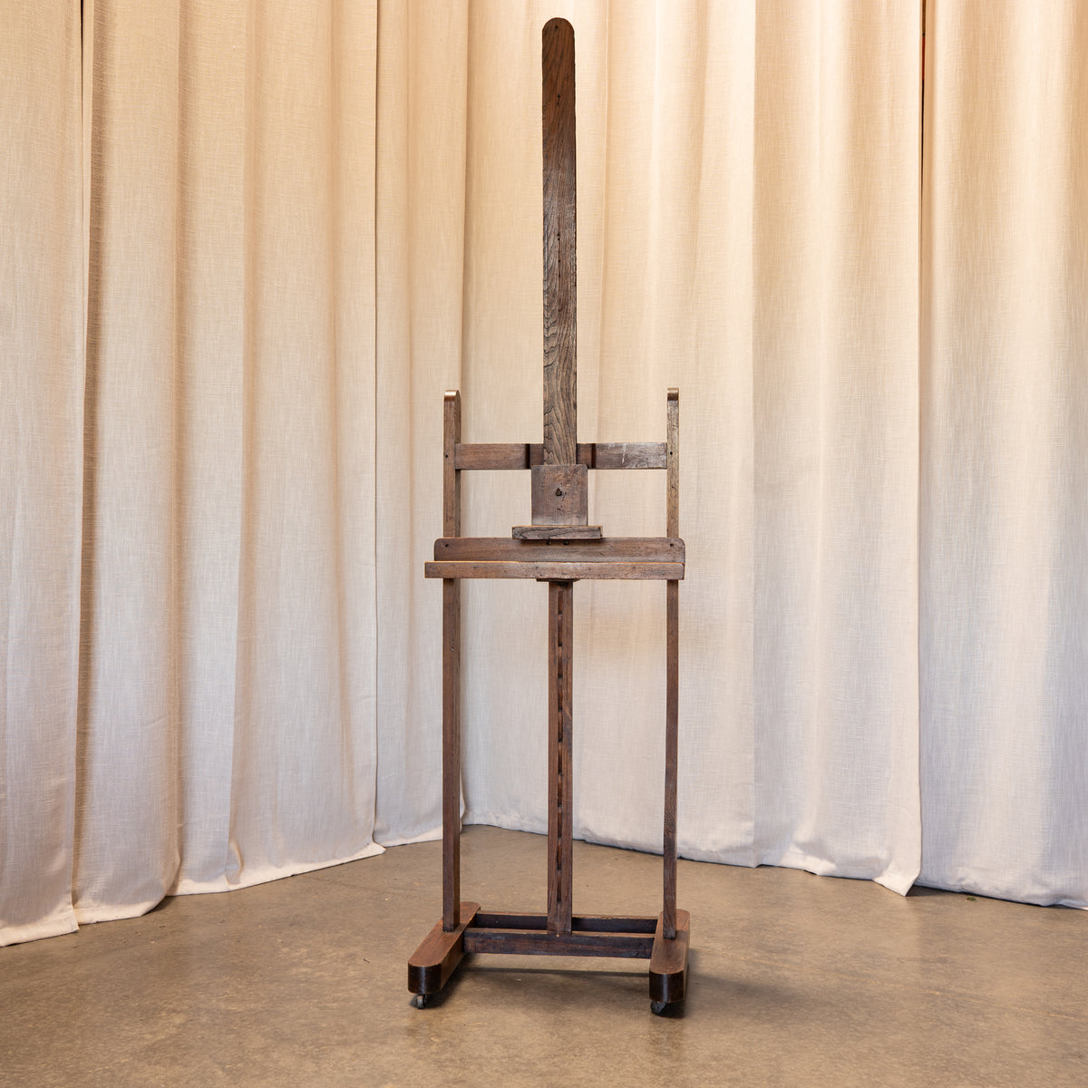 A dark brown wooden artist easel with a tall adjustable arm and footrest, set against a backdrop of draped beige fabric curtains.