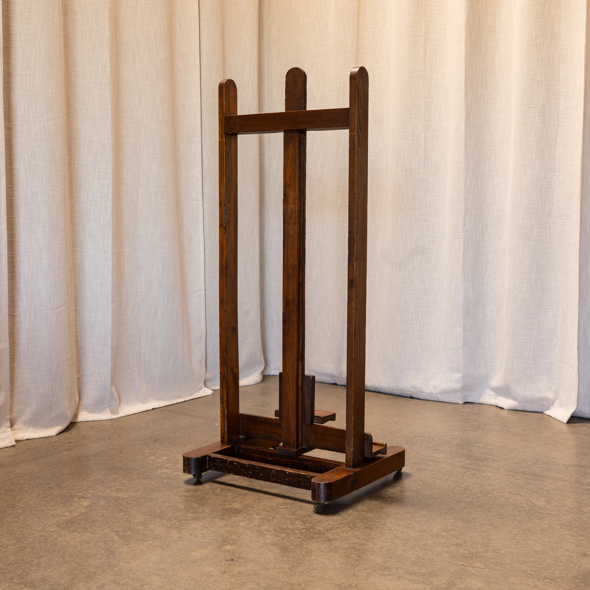 A dark brown wooden easel stand with a wheeled base, positioned against a backdrop of draped off-white curtains and a concrete floor.