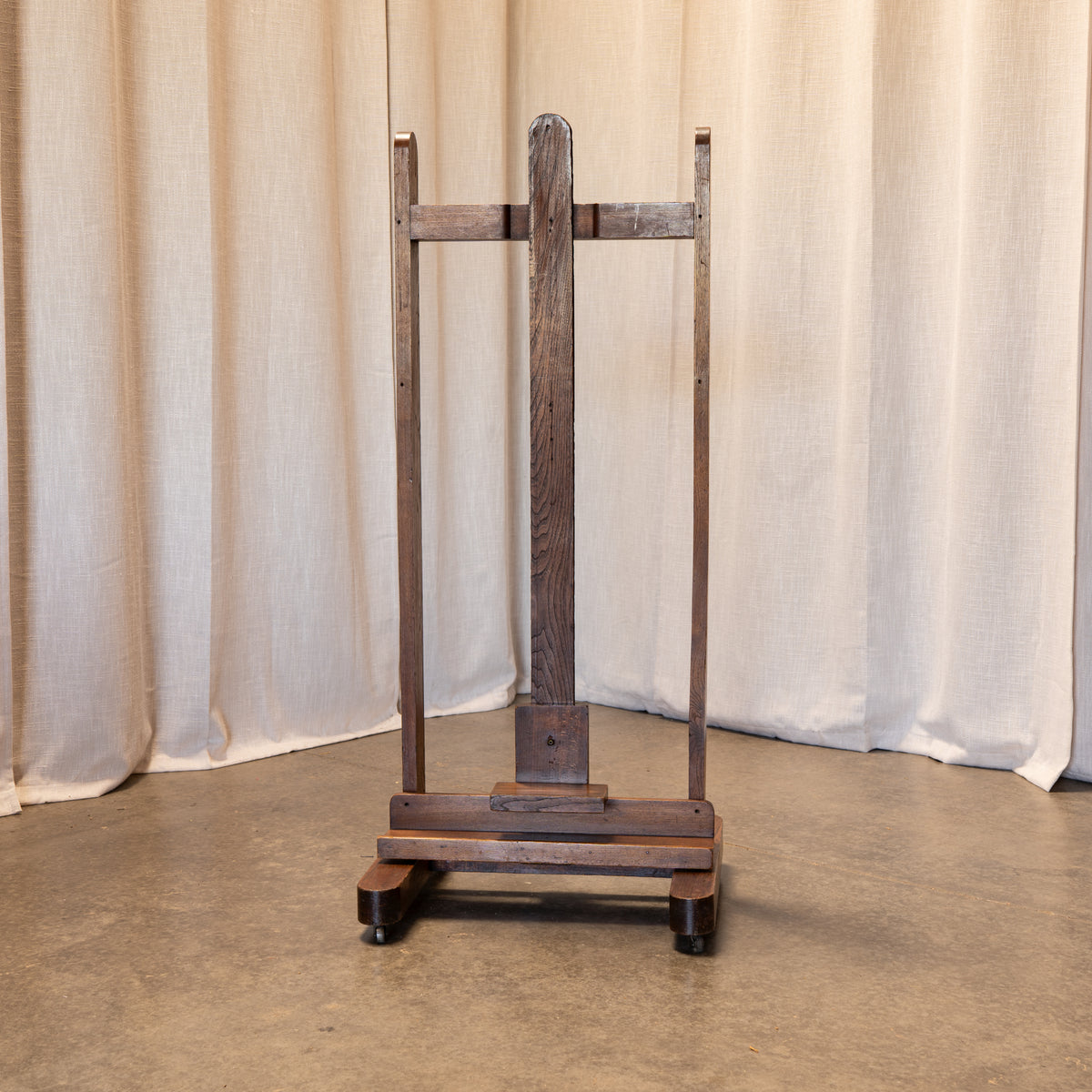 A rustic dark brown wooden easel or stand with a weathered finish, standing on a concrete floor in front of draped beige fabric curtains.