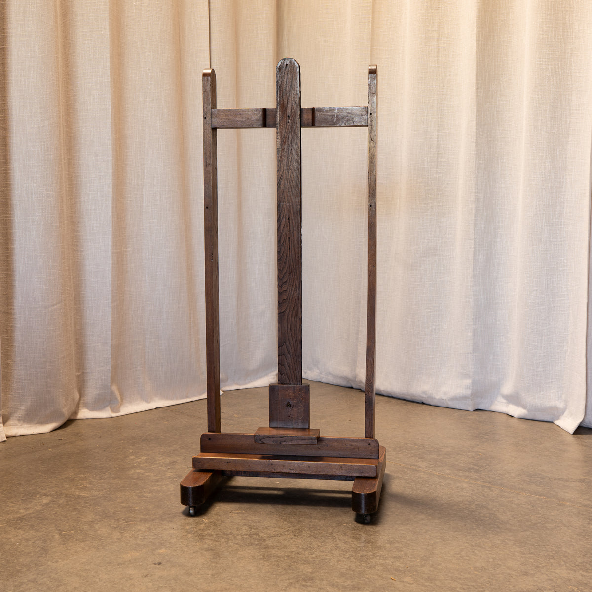 A dark brown wooden display easel or stand with a slatted back panel and a base, photographed indoors against draped beige fabric and a concrete floor.
