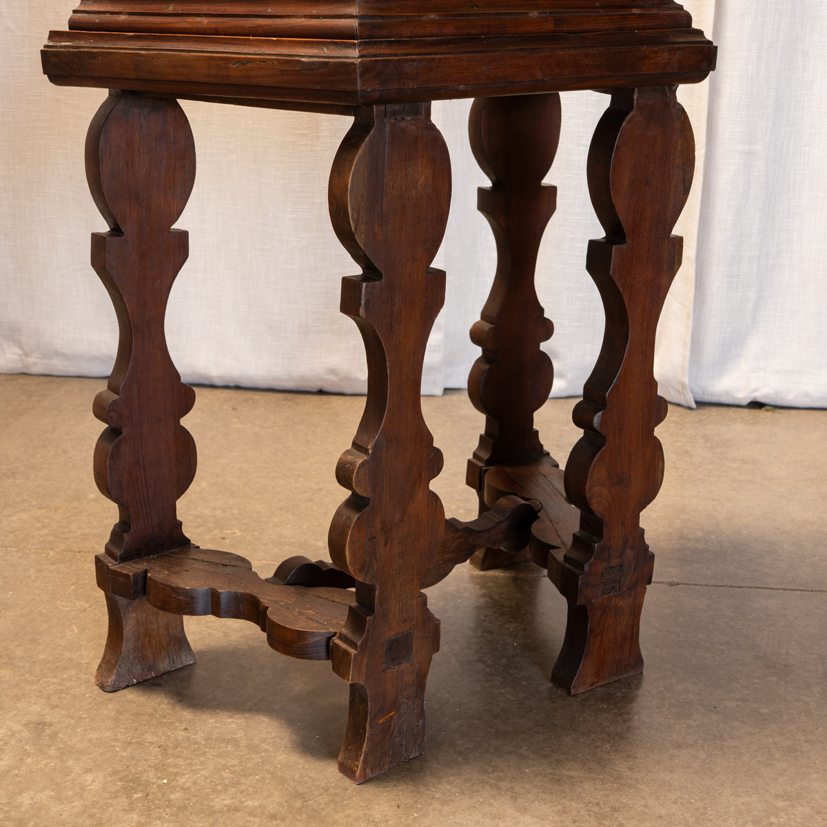 A dark brown wooden side table with ornate turned legs and a stretcher base, photographed against a white draped backdrop and concrete floor.