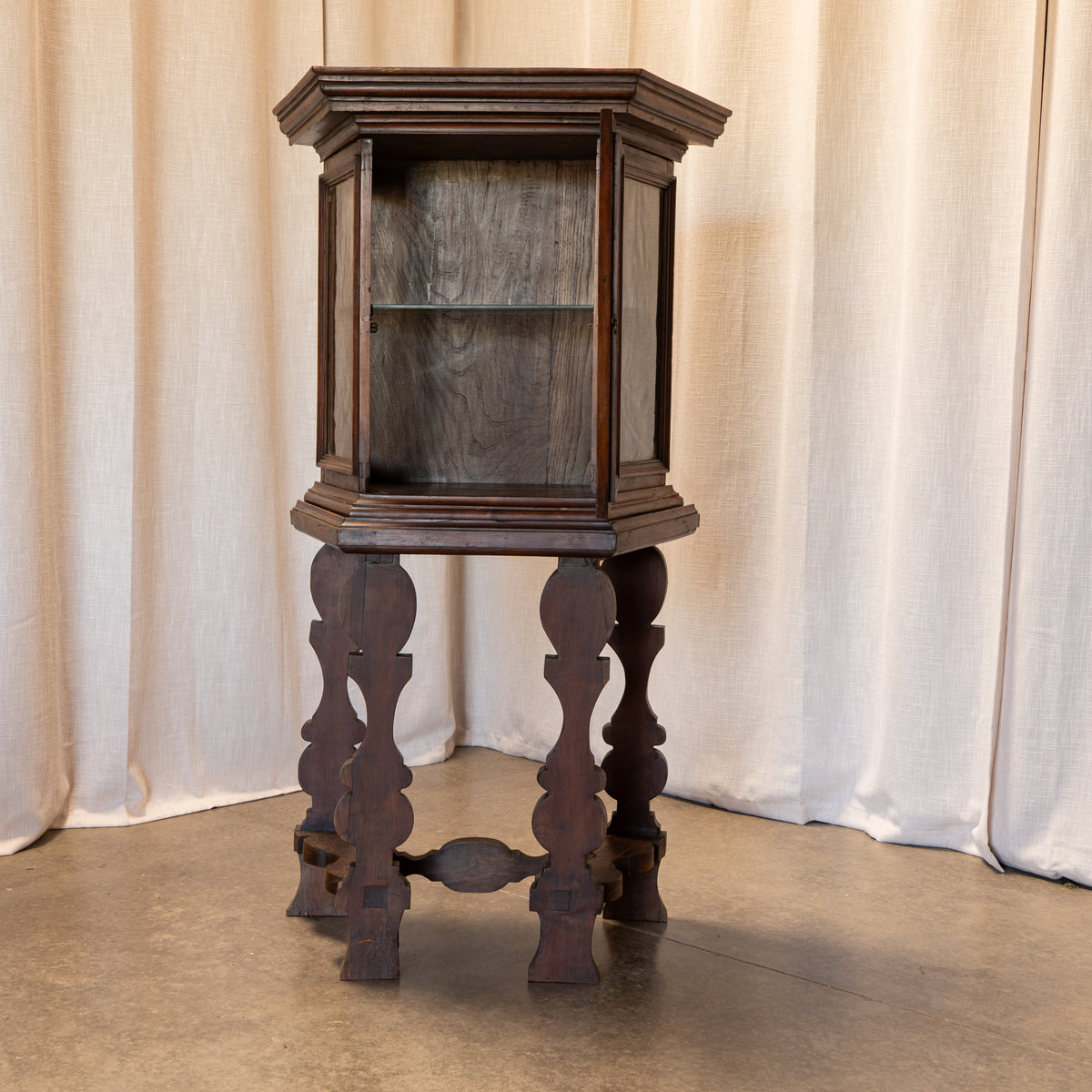 A dark brown wooden display cabinet on a carved pedestal stand, set against a neutral beige curtain backdrop.