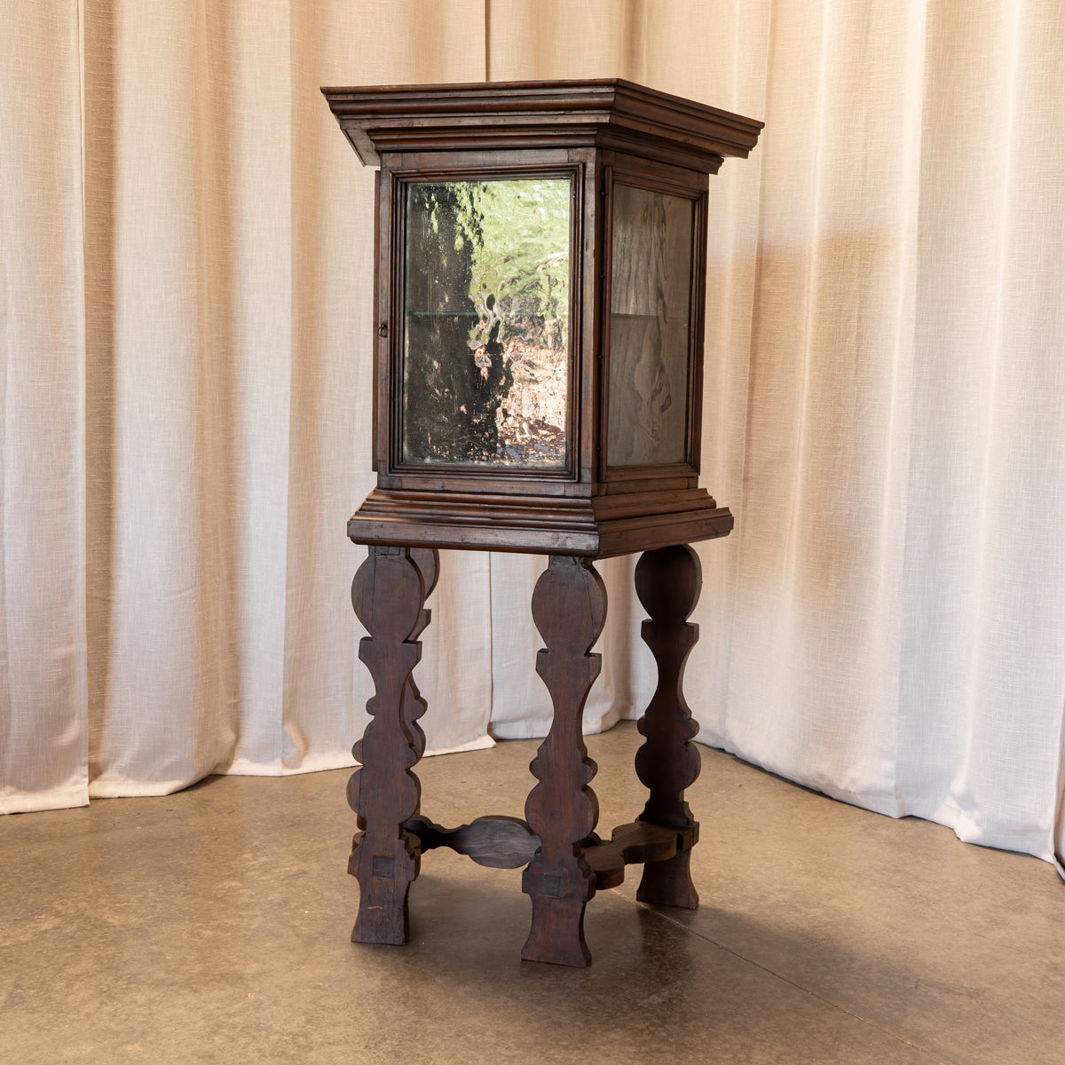 A dark brown wooden display cabinet on a carved pedestal stand, featuring a glass-fronted upper cabinet and a lower cabinet on ornate turned legs, set against a backdrop of beige curtains.