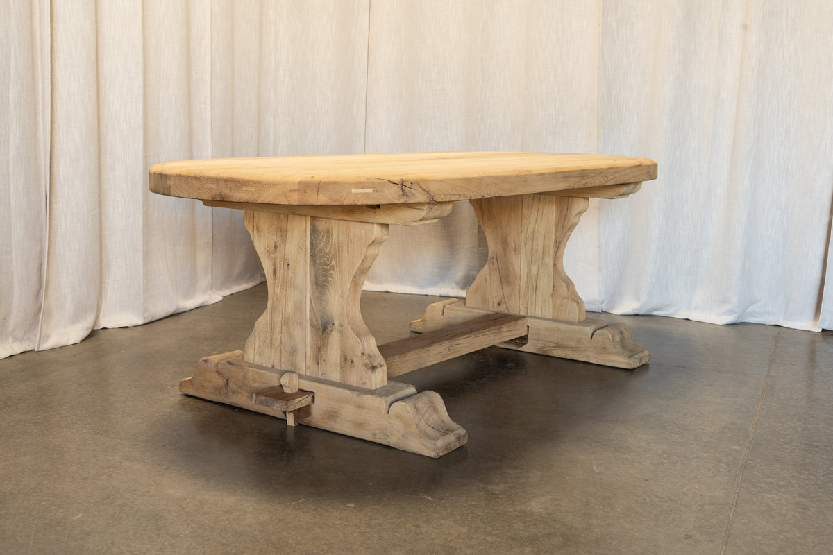 A rustic rectangular wooden dining table with a thick natural wood top and carved trestle-style legs, photographed indoors against a draped white fabric backdrop and concrete floor.