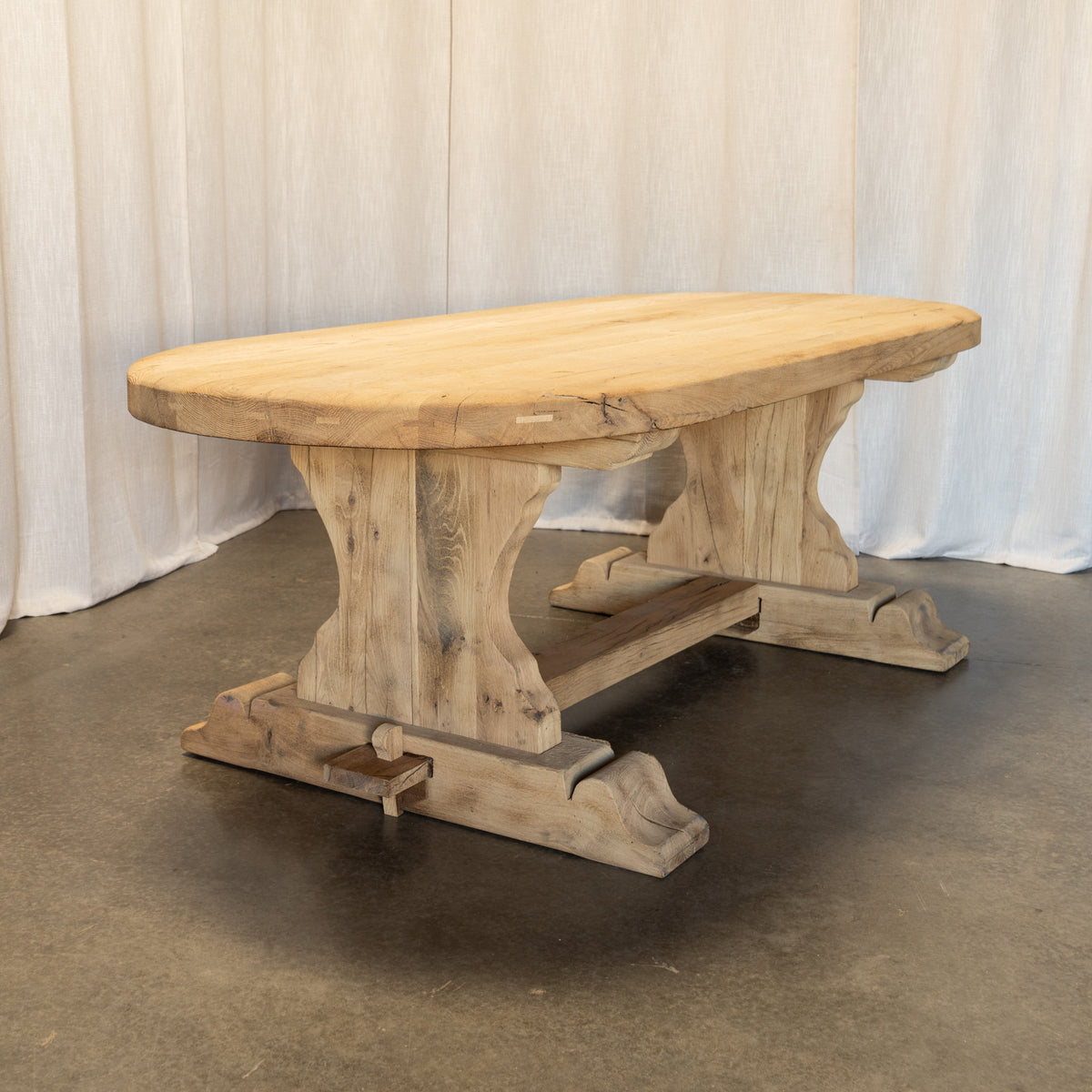 A rustic oval wooden dining table with a natural light wood top and weathered trestle-style legs, photographed in front of a white draped backdrop on a gray concrete floor.