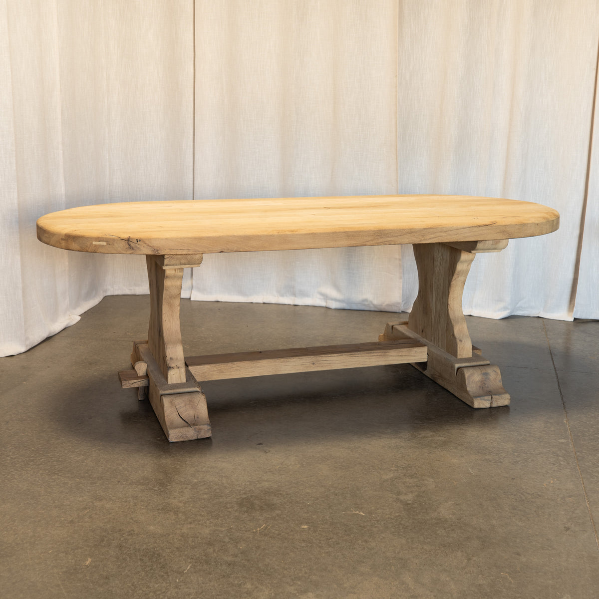 A rustic oval wooden dining table with a light natural top and weathered gray-brown trestle-style legs, set against a draped fabric backdrop.