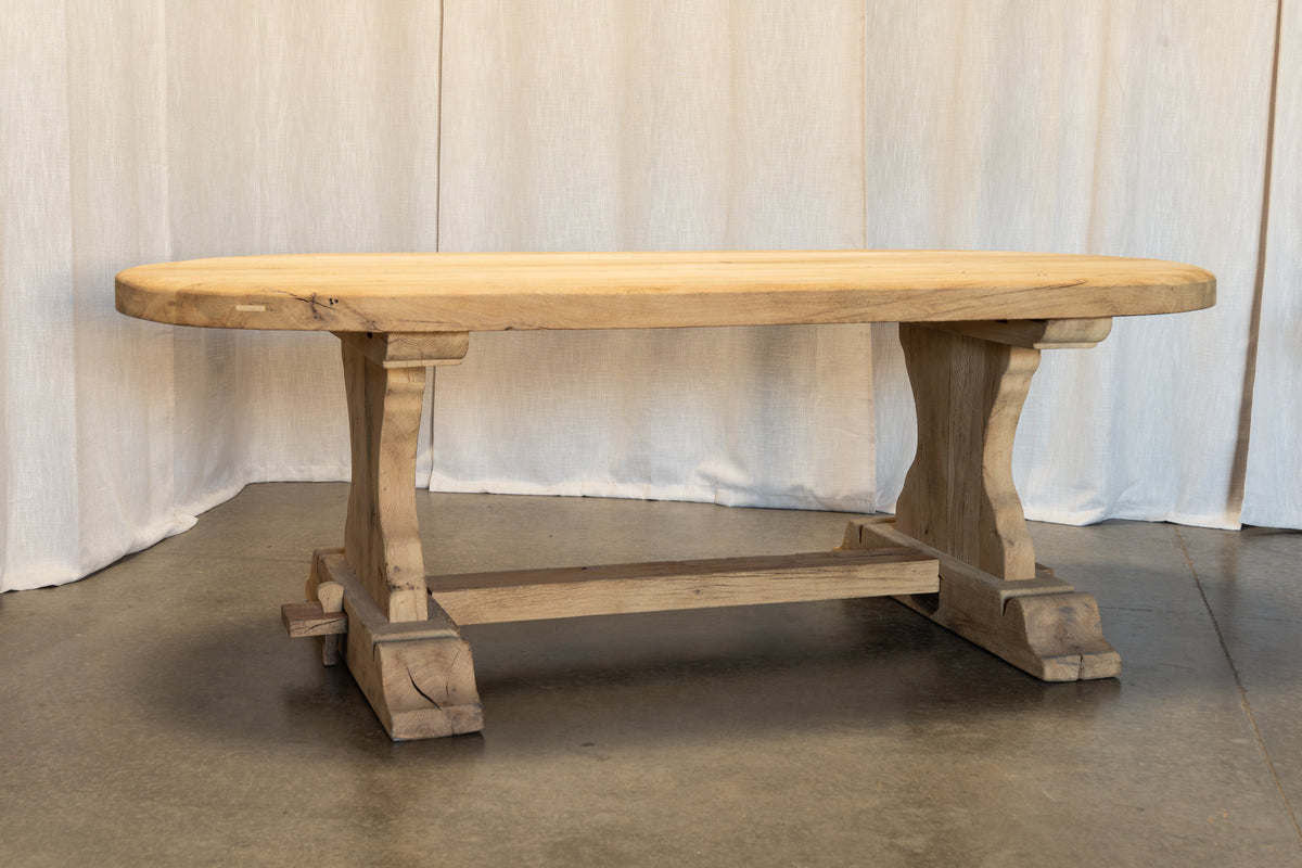 A rustic light oak wood trestle table with a thick natural tabletop and sturdy block-style legs, set against a draped white fabric backdrop.